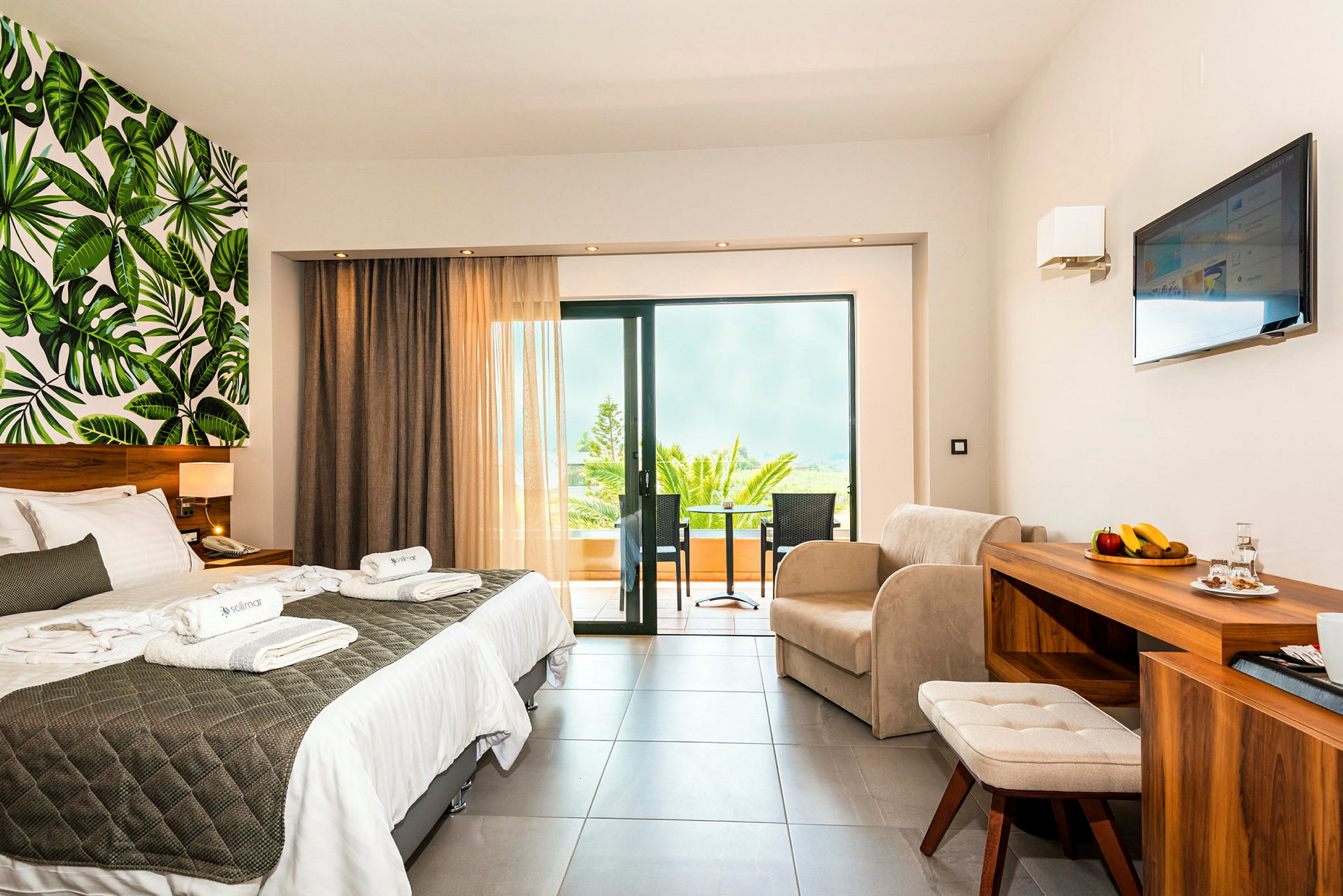 Ett hotellrum med dubbelsäng, sittgrupp, balkong och väggdesign med bladmotiv. (AI-genererad text)