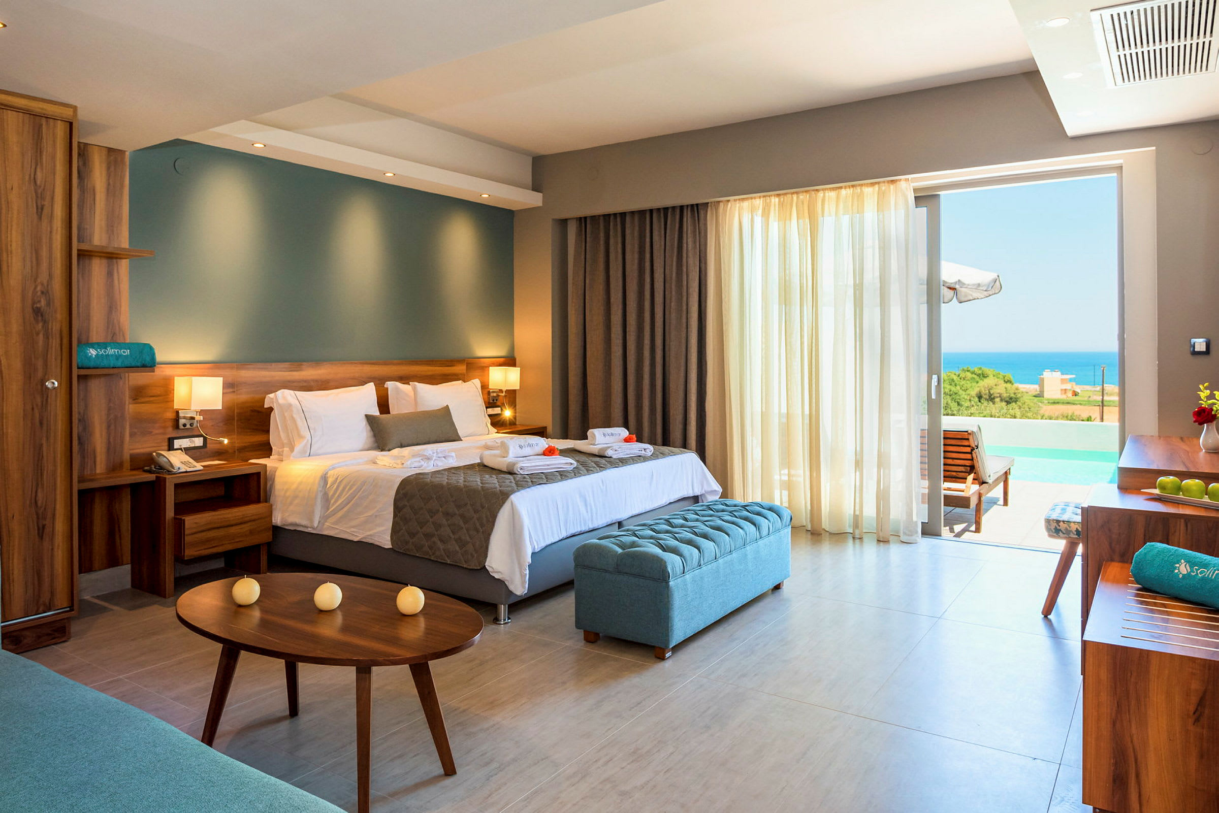 Hotellrum med stor säng, sittmöbler och utsikt över havet genom glasdörrar. (AI-genererad text)