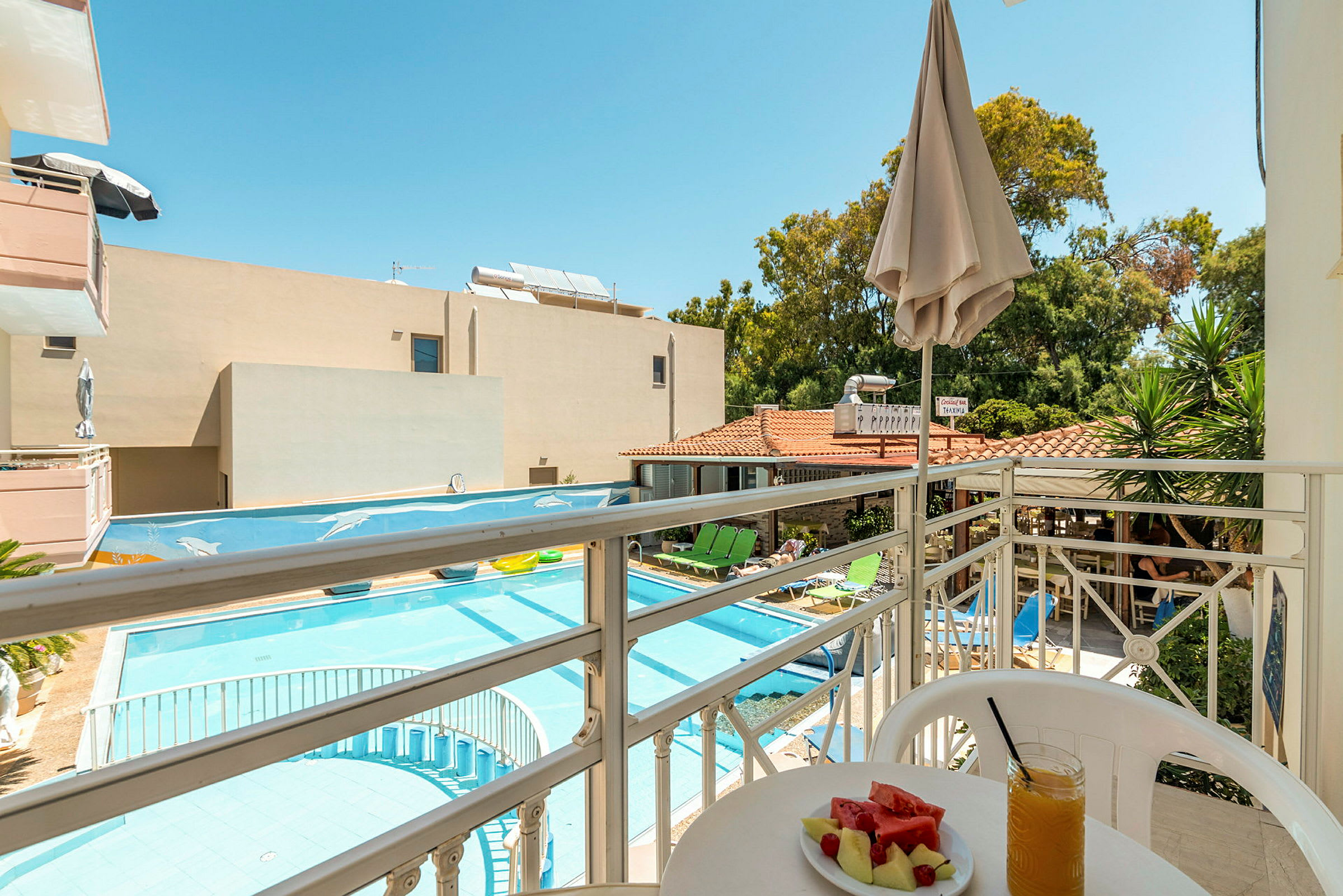 Utsikt från en balkong mot en pool och hotellområde med solstolar, parasoller och grönska. På balkongen finns ett bord med en tallrik melon och ett glas juice. (AI-genererad text)