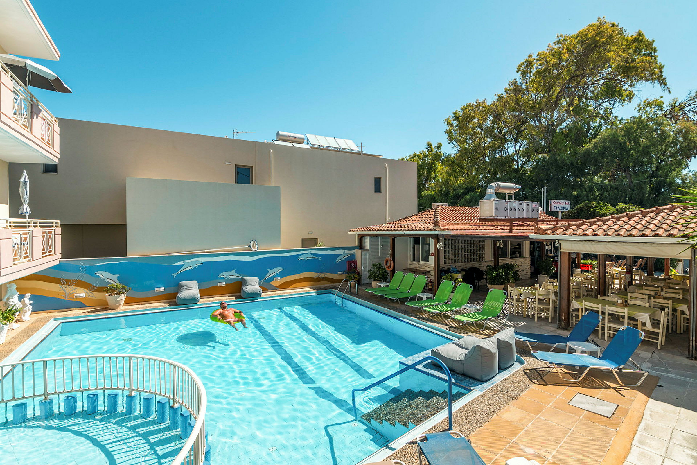 Bild av en pool vid ett hotell med solstolar och en muralmålning på väggen. En person flyter i vattnet. (AI-genererad text)