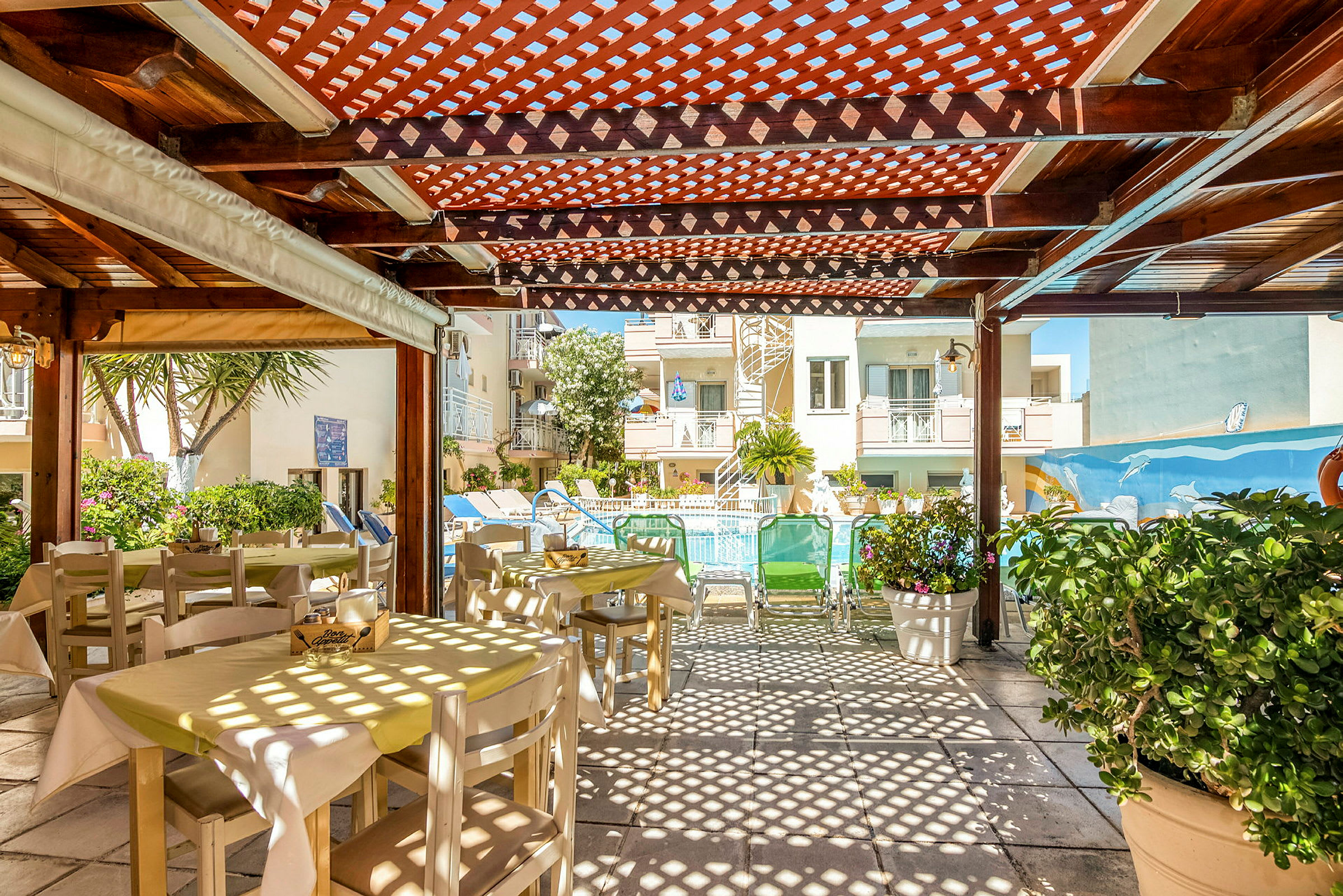 Bild av en utomhus matplats med bord och stolar under en träpergola, med utsikt mot en pool och byggnader i bakgrunden. (AI-genererad text)