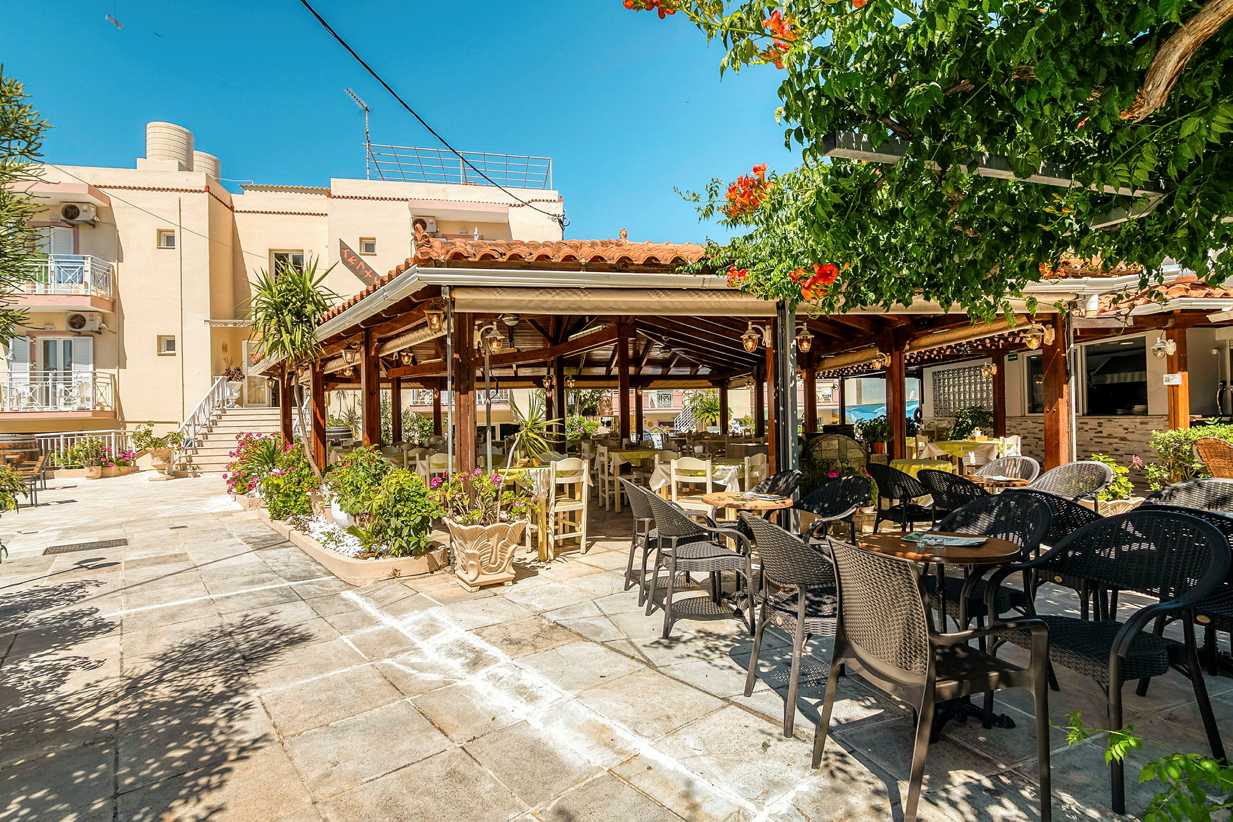 En utomhusrestaurang med bord och stolar under en pergola, omgiven av gröna växter. (AI-genererad text)