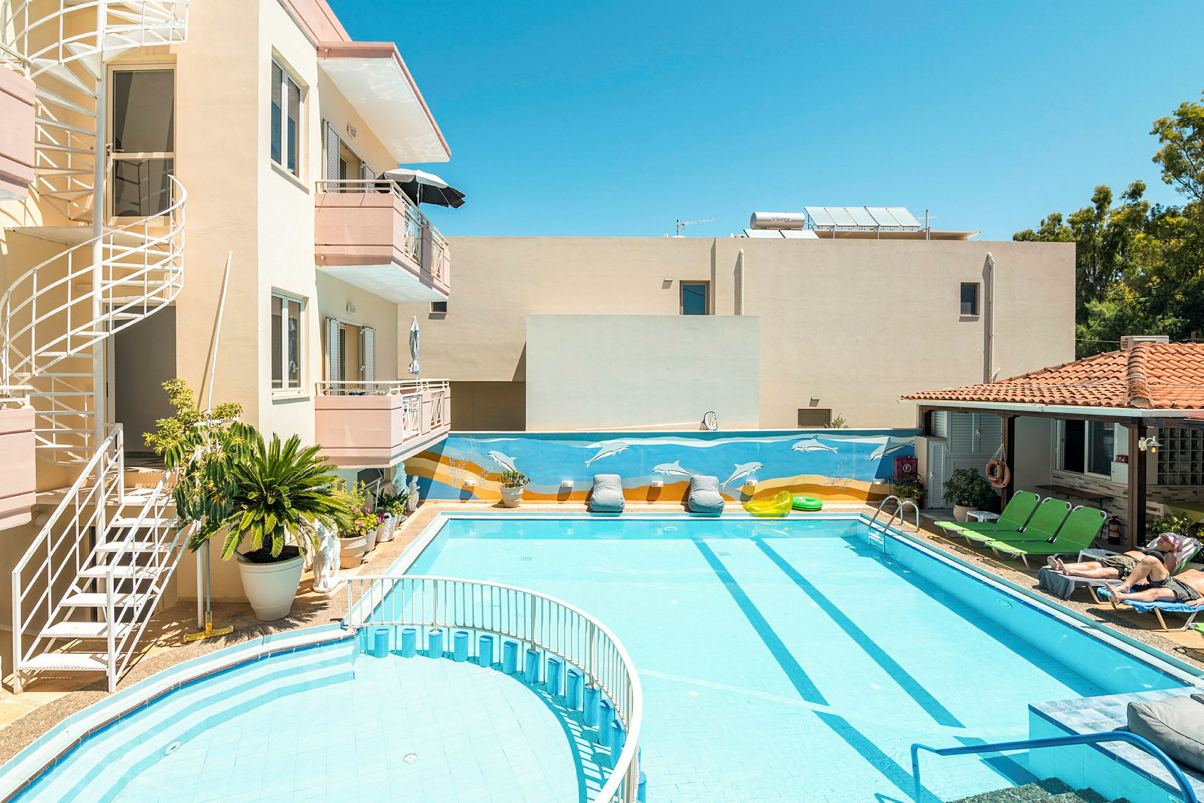 En byggnad med balkonger och spiraltrappa vid en pool. Poolområdet har solsängar och dekorationer, inklusive en väggmålning med delfiner. (AI-genererad text)