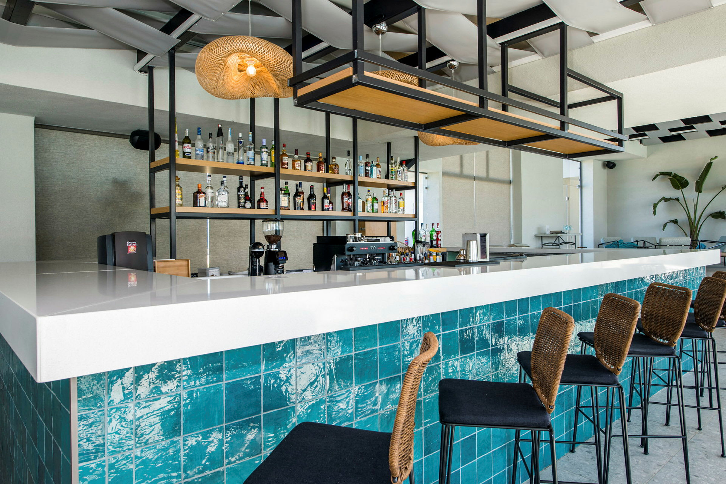 En modern bar med en vit bardisk och blå kakel, hyllor med alkoholflaskor i bakgrunden och höga stolar med rottingryggar. (AI-genererad text)