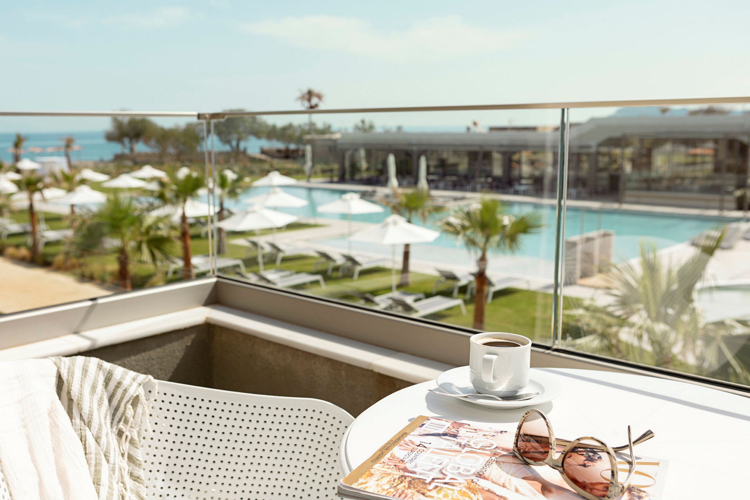 Utsikt över en pool och trädgård från en balkong med ett bord som har en kaffekopp, en tidning och solglasögon. (AI-genererad text)