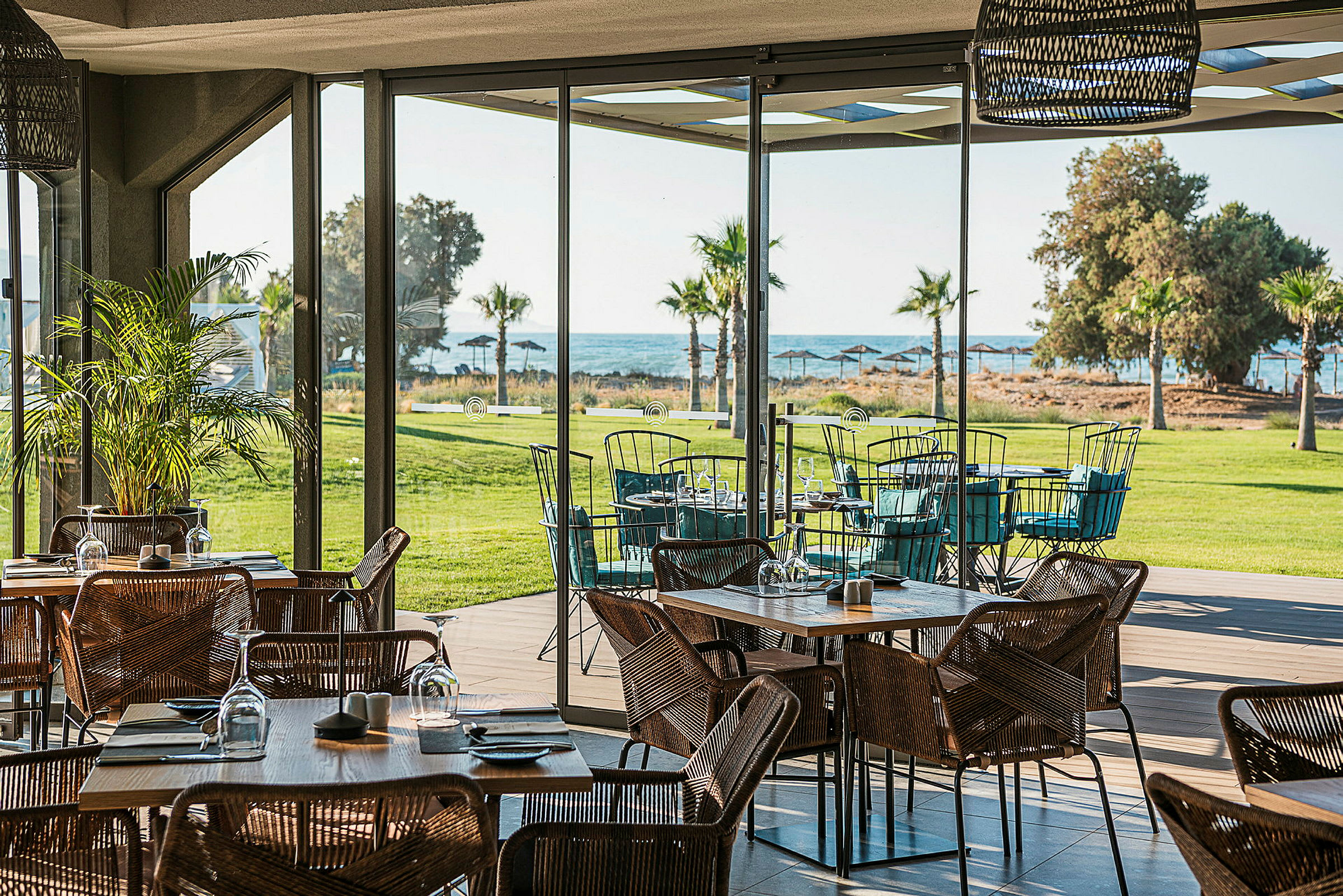 En restaurang med glasväggar och utsikt över en grön gräsmatta, palmer och havet. (AI-genererad text)