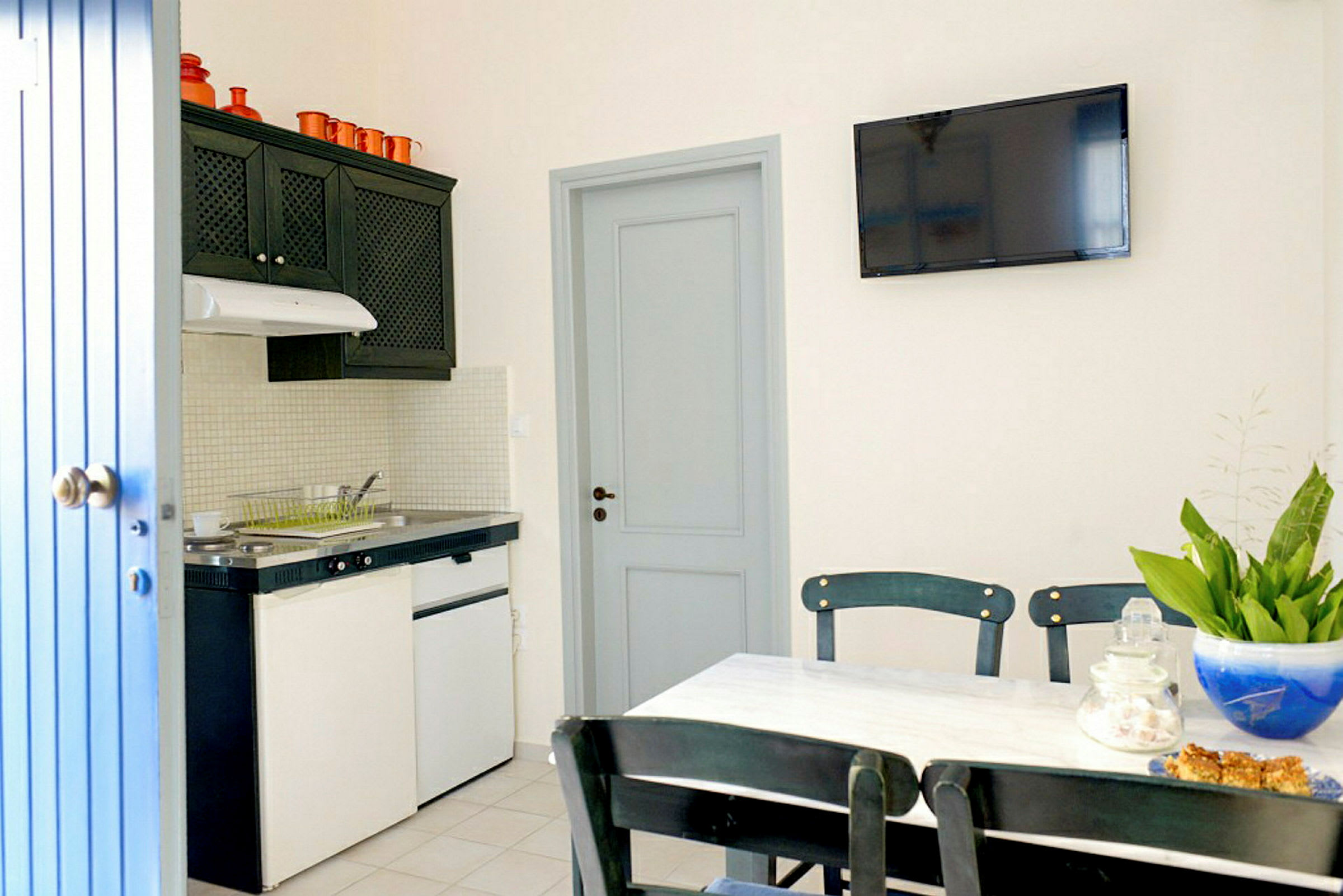 Ett kök med spiselkrans, kylskåp och diskho. Ett matbord med en blå skål och gröna växter i förgrunden. (AI-genererad text)