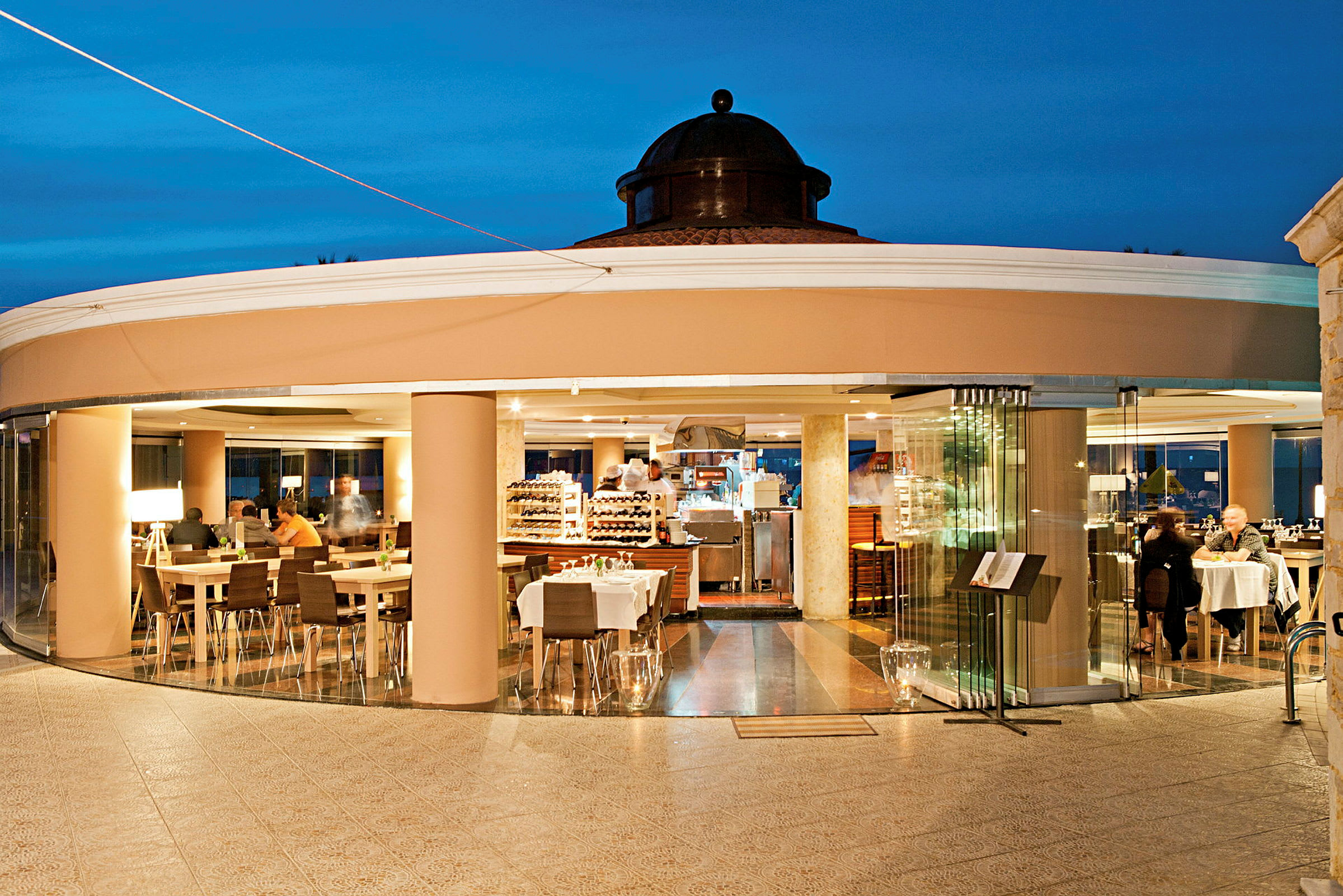 En restaurang med glasväggar och bord belägna inomhus, sett från en utomhusterrass. (AI-genererad text)