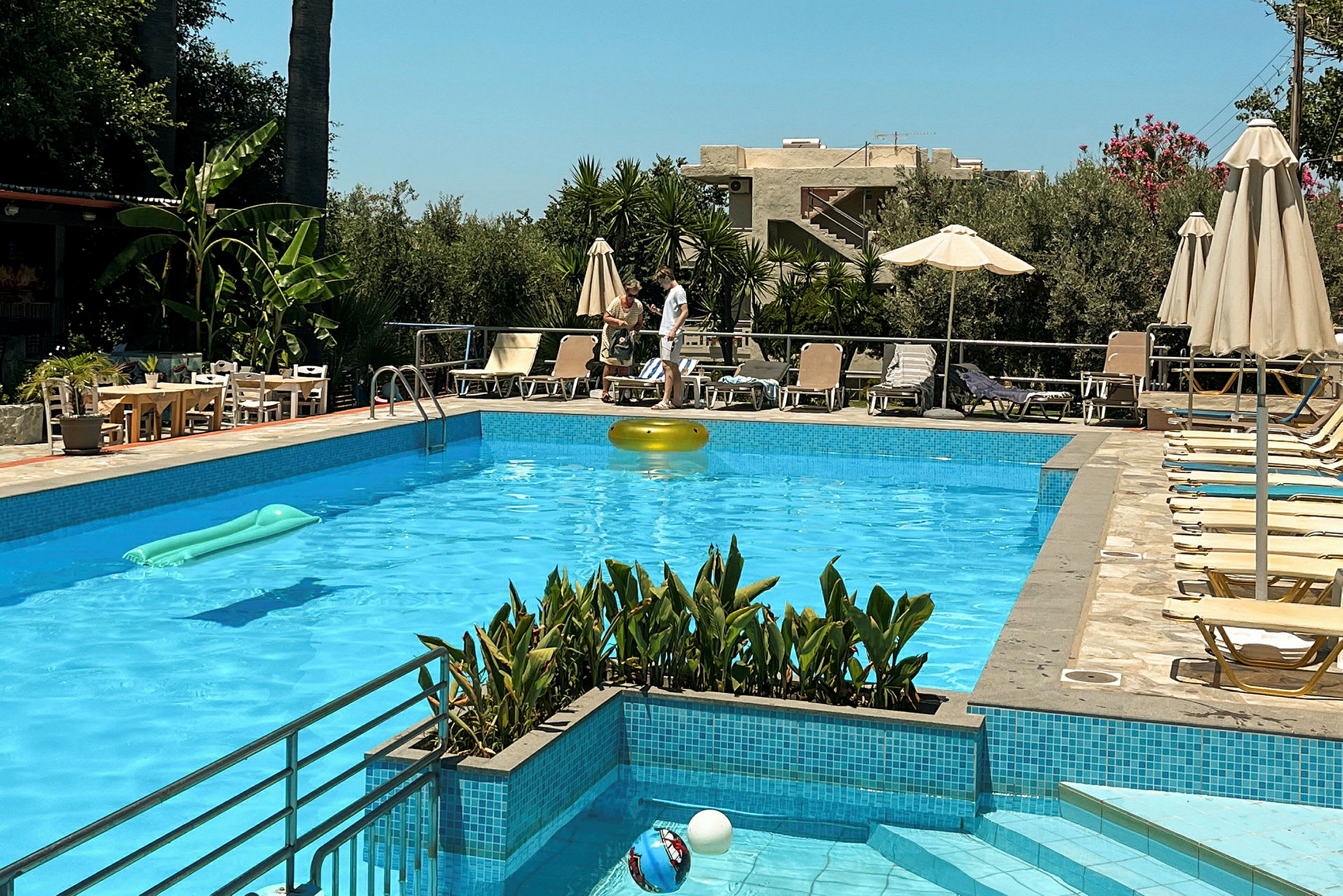 En swimmingpool med solsängar och parasoller runtomkring. I bakgrunden syns växtlighet och byggnader. (AI-genererad text)