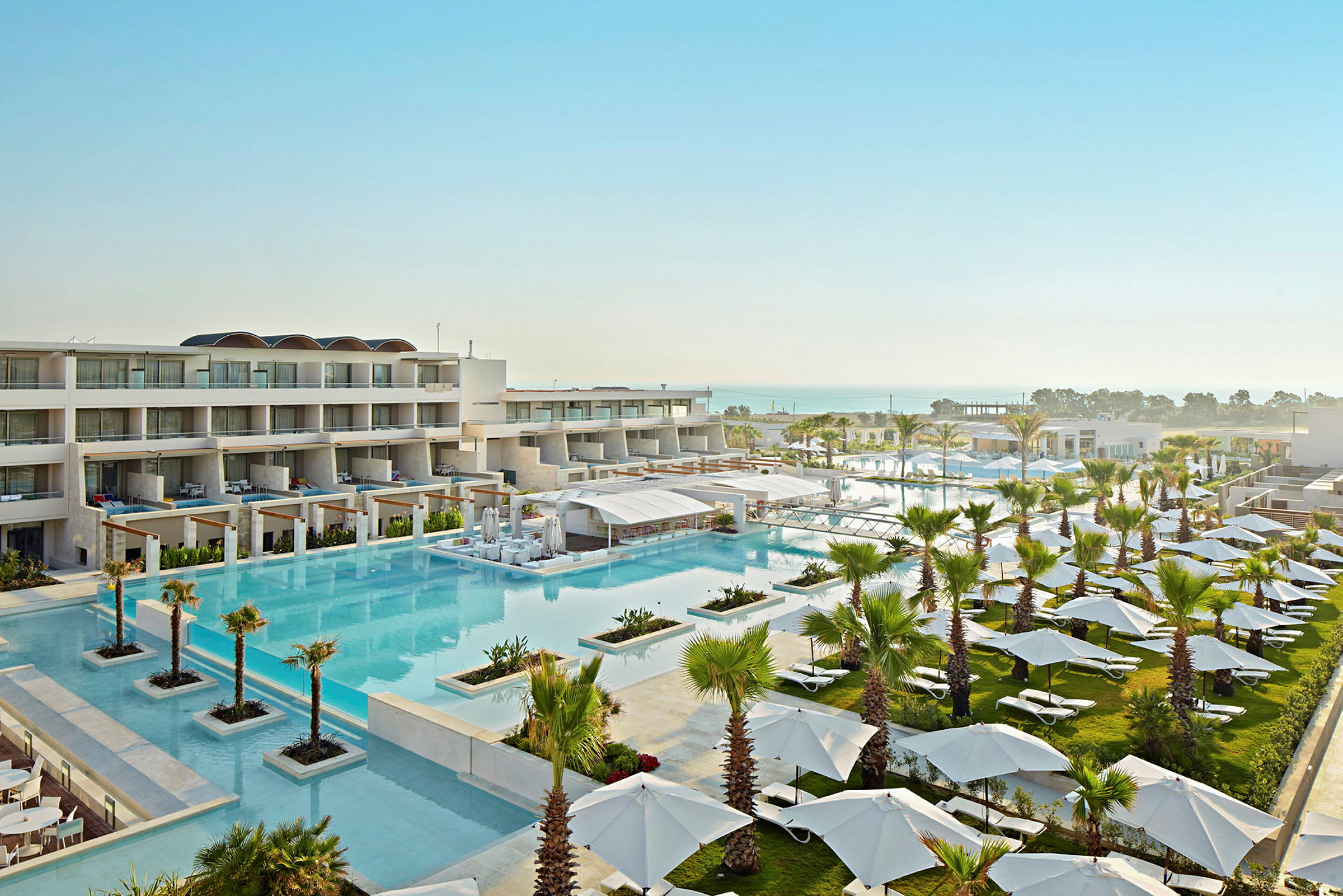 Ett hotellområde med flera byggnader, en stor pool och omgivande gröna områden fyllda med parasoller och solstolar. I bakgrunden syns havet. (AI-genererad text)