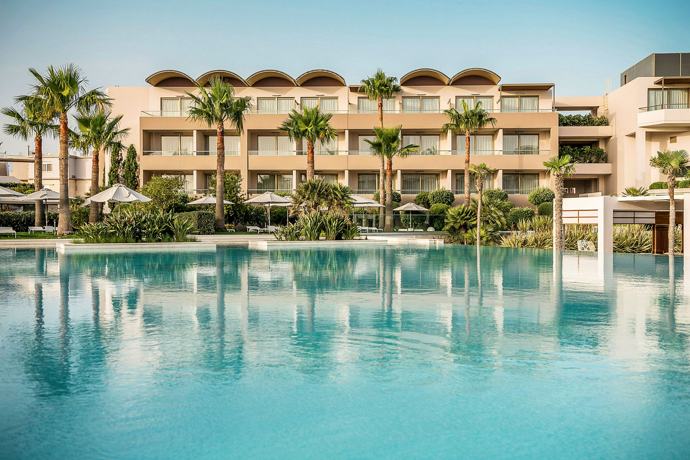 Ett stort poolområde med klarblått vatten framför ett modernt, flervåningshotell med palmer och grönska. (AI-genererad text)