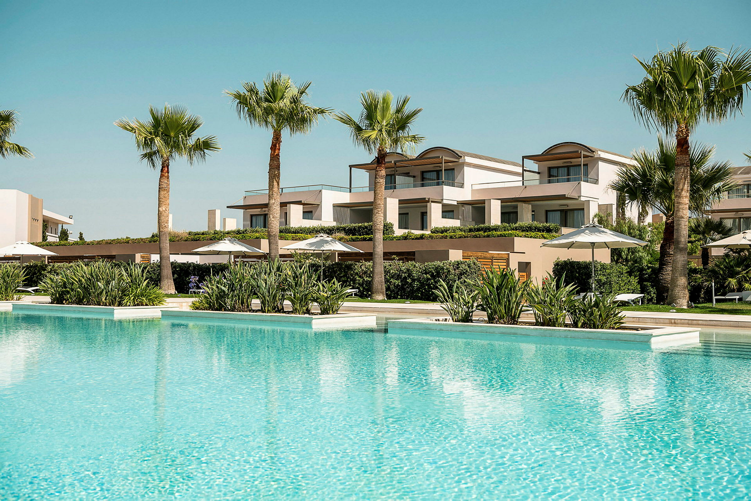 På bilden syns en stor swimmingpool med klart vatten, omgiven av palmer och moderna hotellbyggnader i bakgrunden. (AI-genererad text)