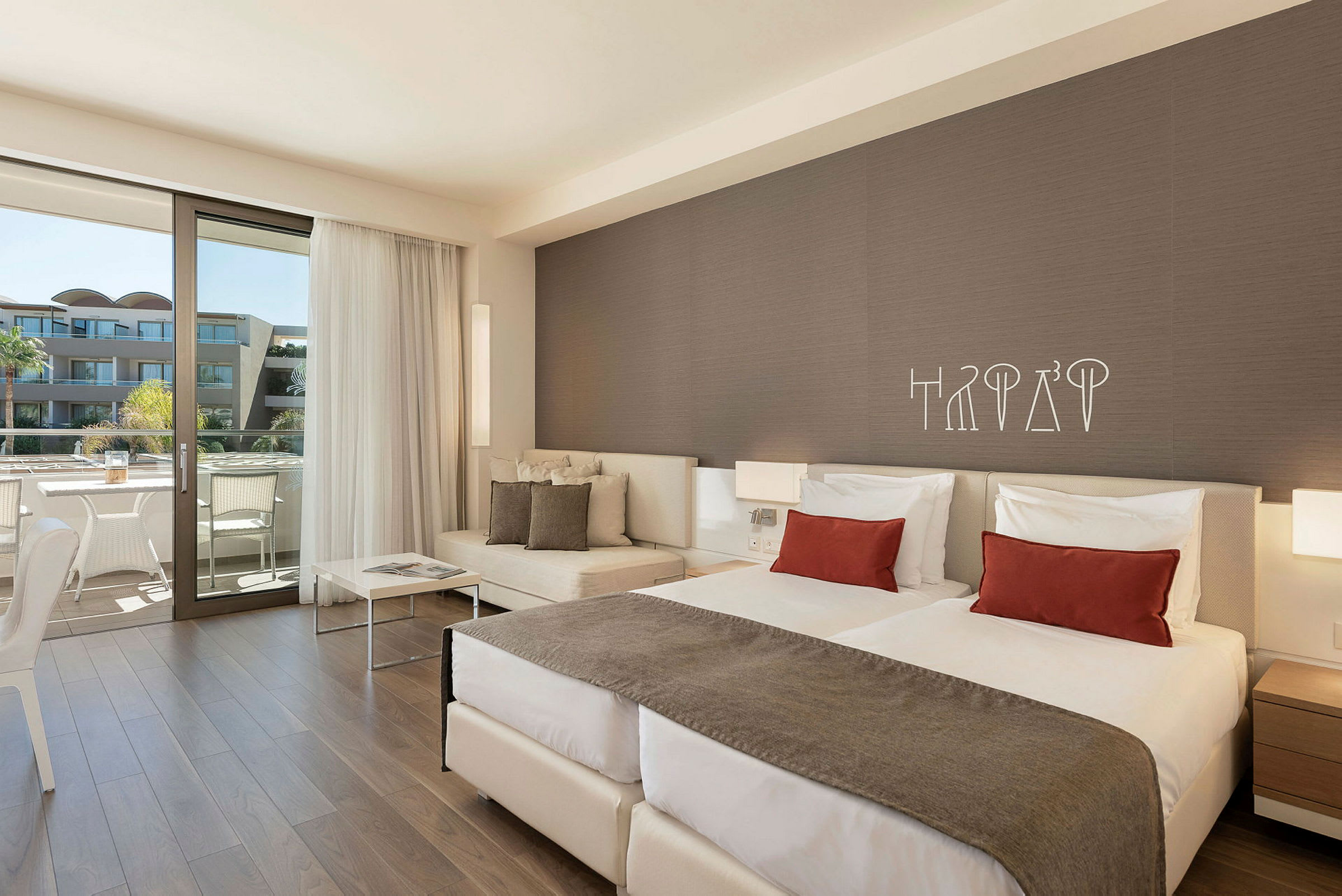 Ett hotellrum med två sängar, en soffa och stor balkong med utsikt mot byggnader och palmer. (AI-genererad text)