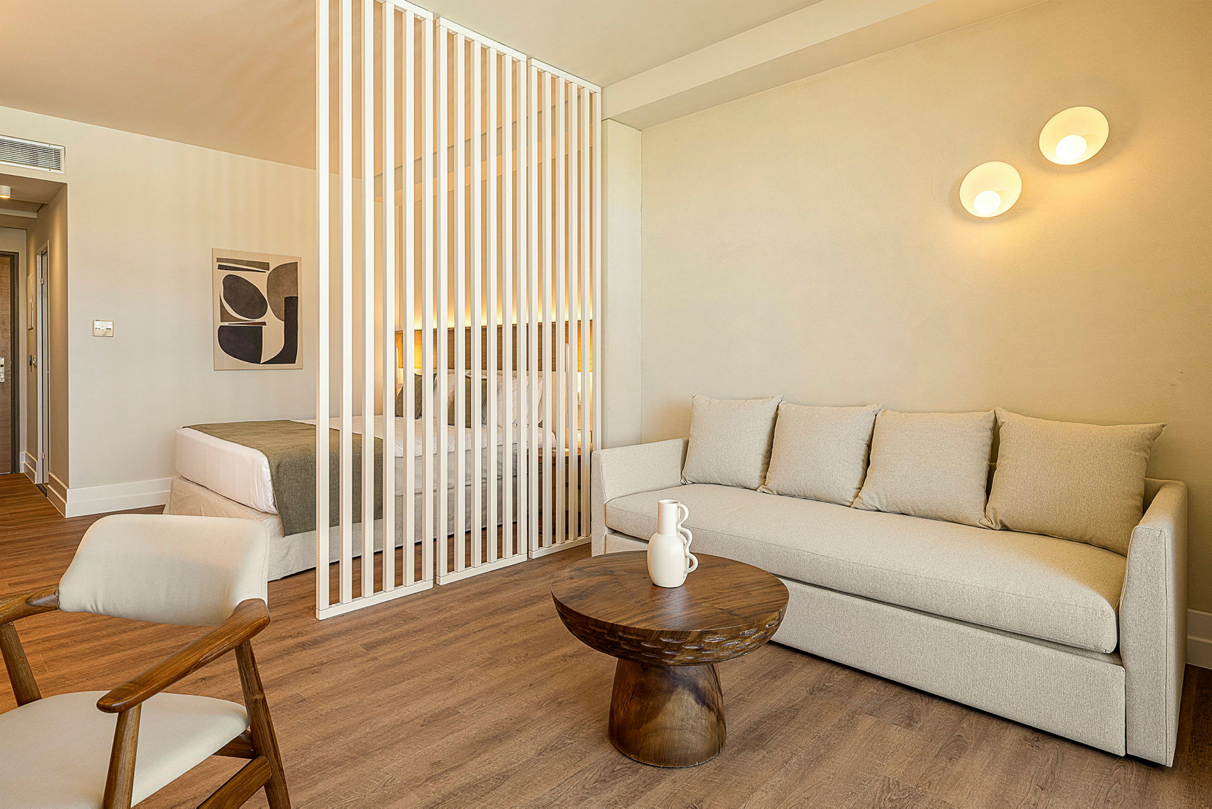Ett rum med en sittgrupp bestående av en beige soffa och ett litet runt träbord framför. I bakgrunden finns en dubbelsäng avskild med vita vertikala ribbor. (AI-genererad text)