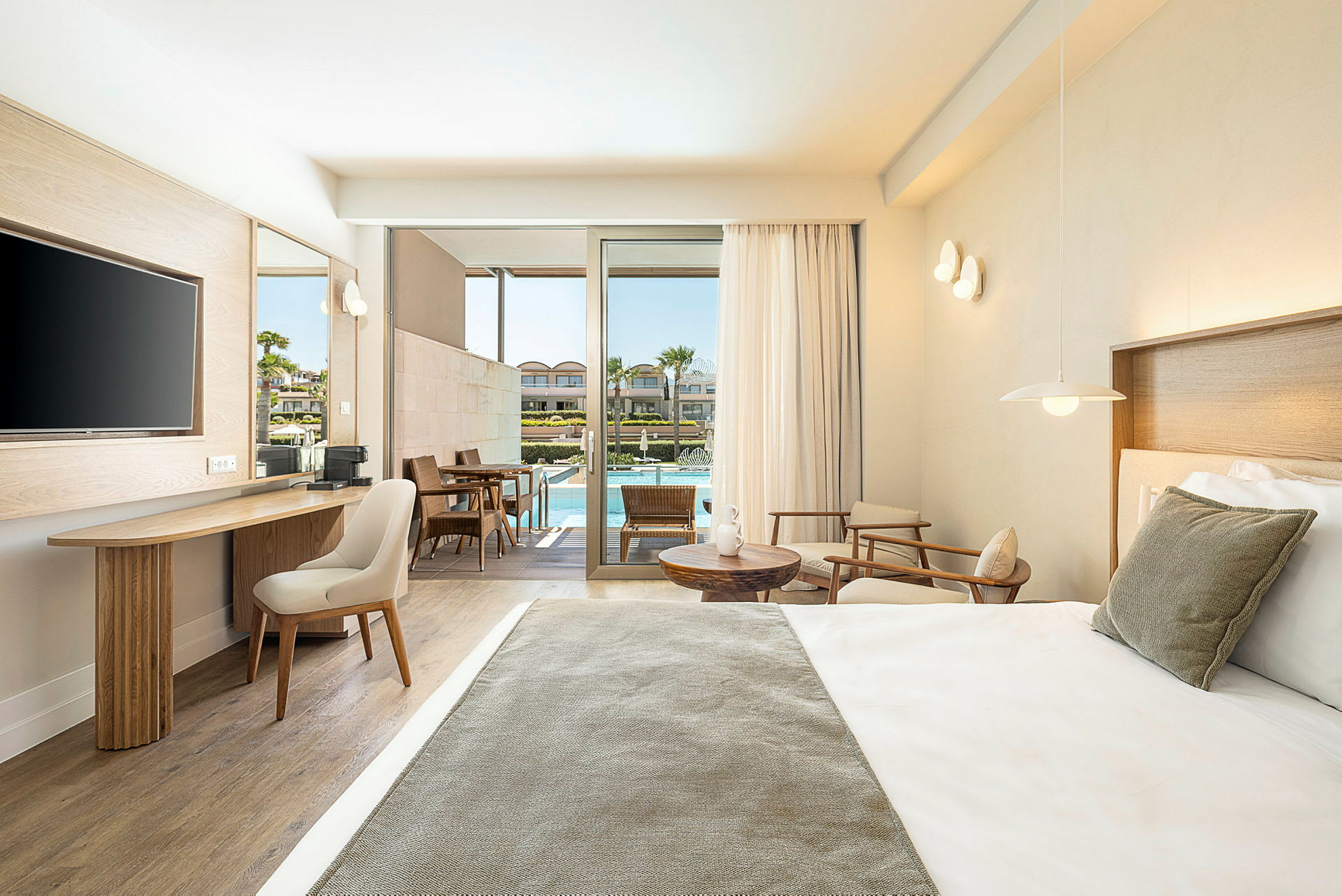Ett modernt hotellrum med en bäddad säng, sittgrupp och skrivbord. Rummet har stora glasdörrar ut mot en uteplats med bord, stolar och tillgång till en pool. (AI-genererad text)