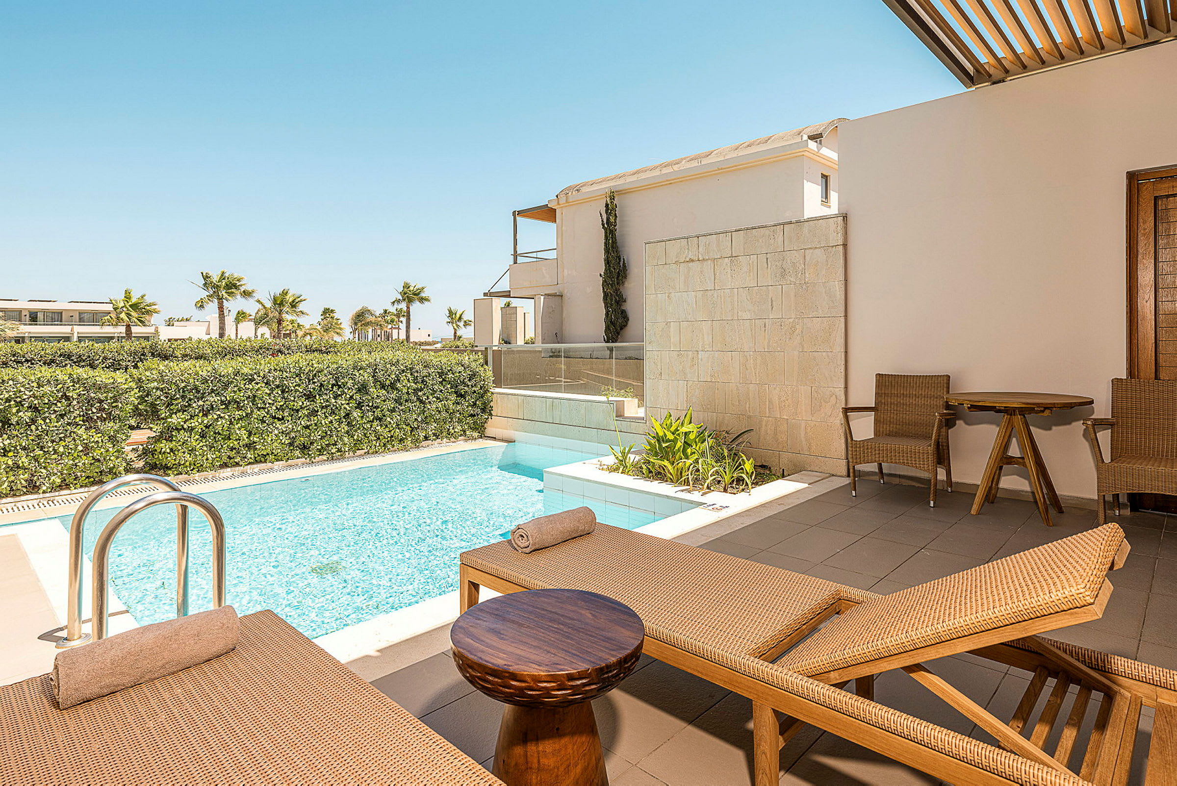 Bild föreställer en terrass med solstolar, ett litet bord och två stolar bredvid en privat pool. I bakgrunden syns grönskande buskar och palmer. (AI-genererad text)