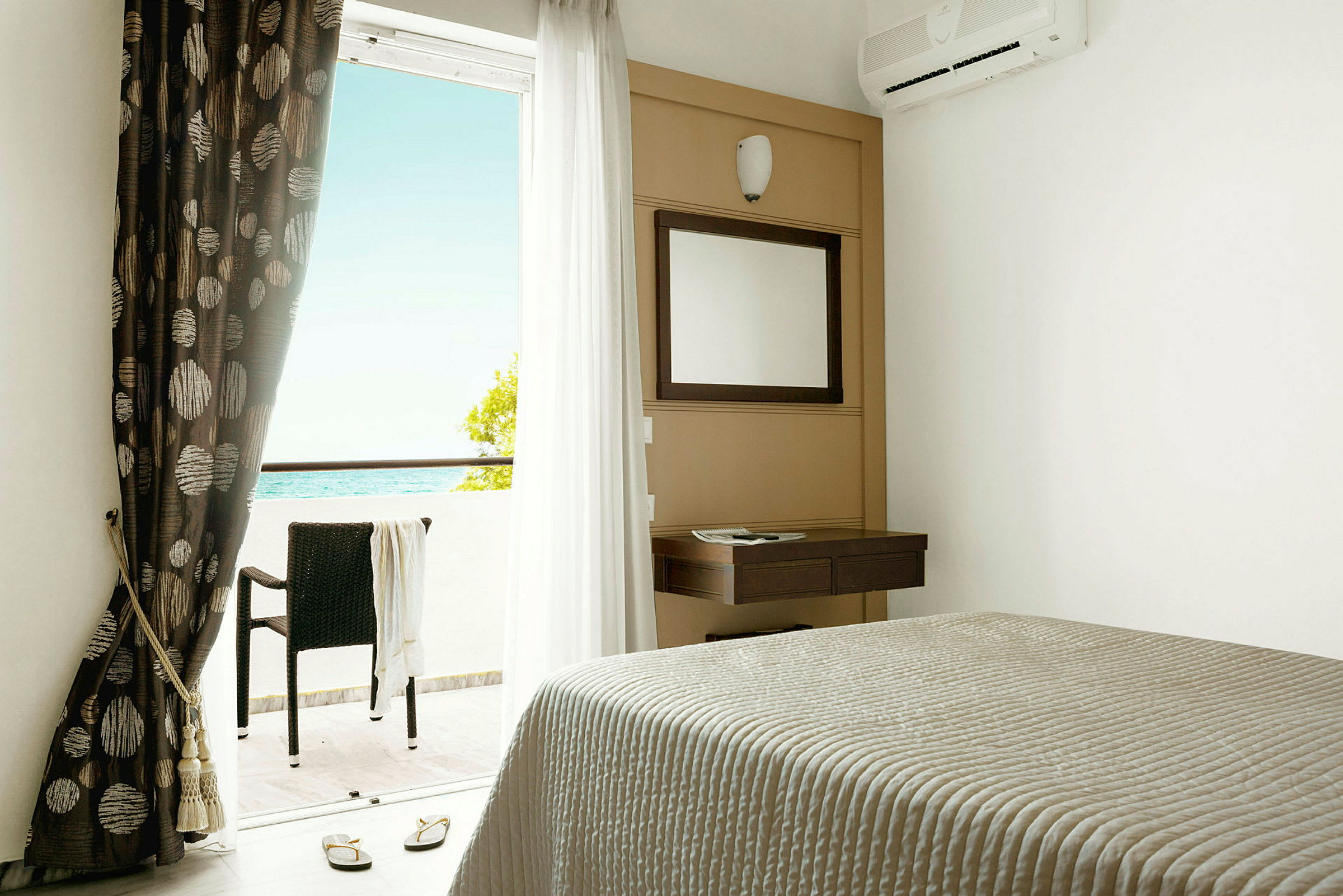 Hotellrum med säng, spegel, gardiner och balkong med utsikt mot havet. (AI-genererad text)
