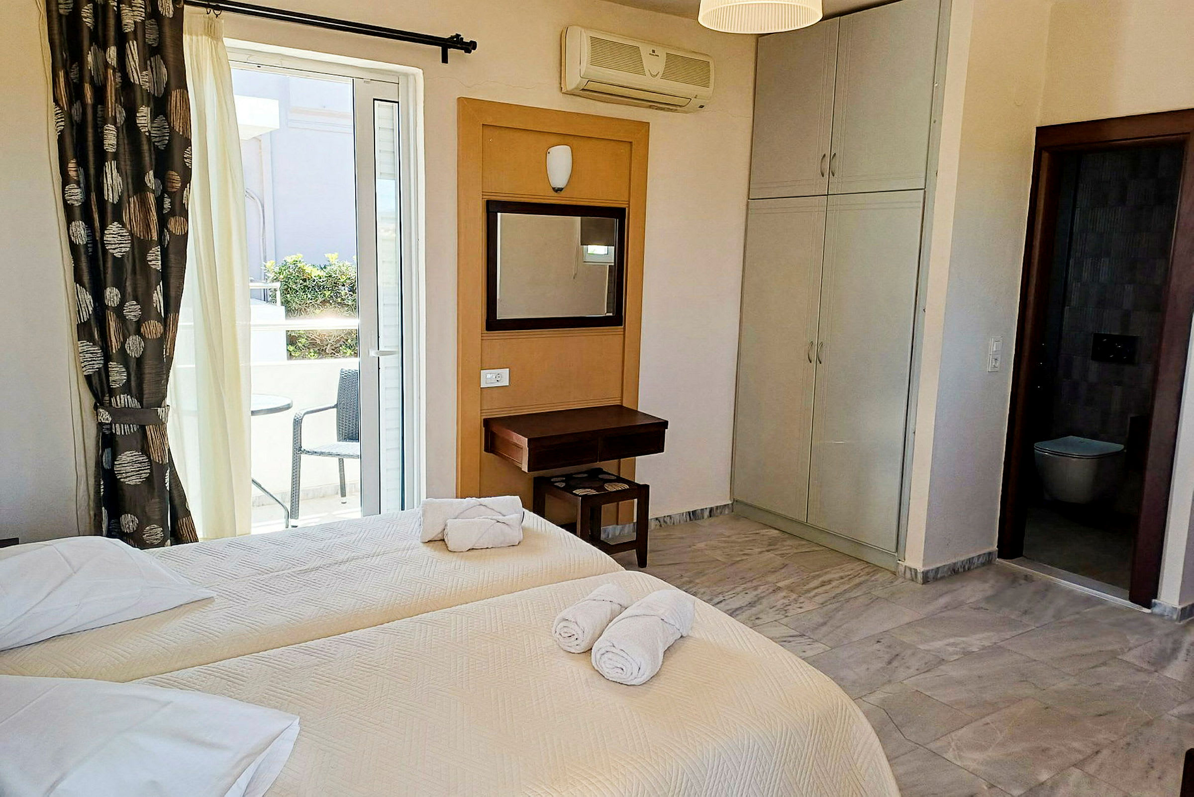 Hotel rum med två enkelsängar, ett skrivbord, och utsikt mot en balkong med sittplats. (AI-genererad text)