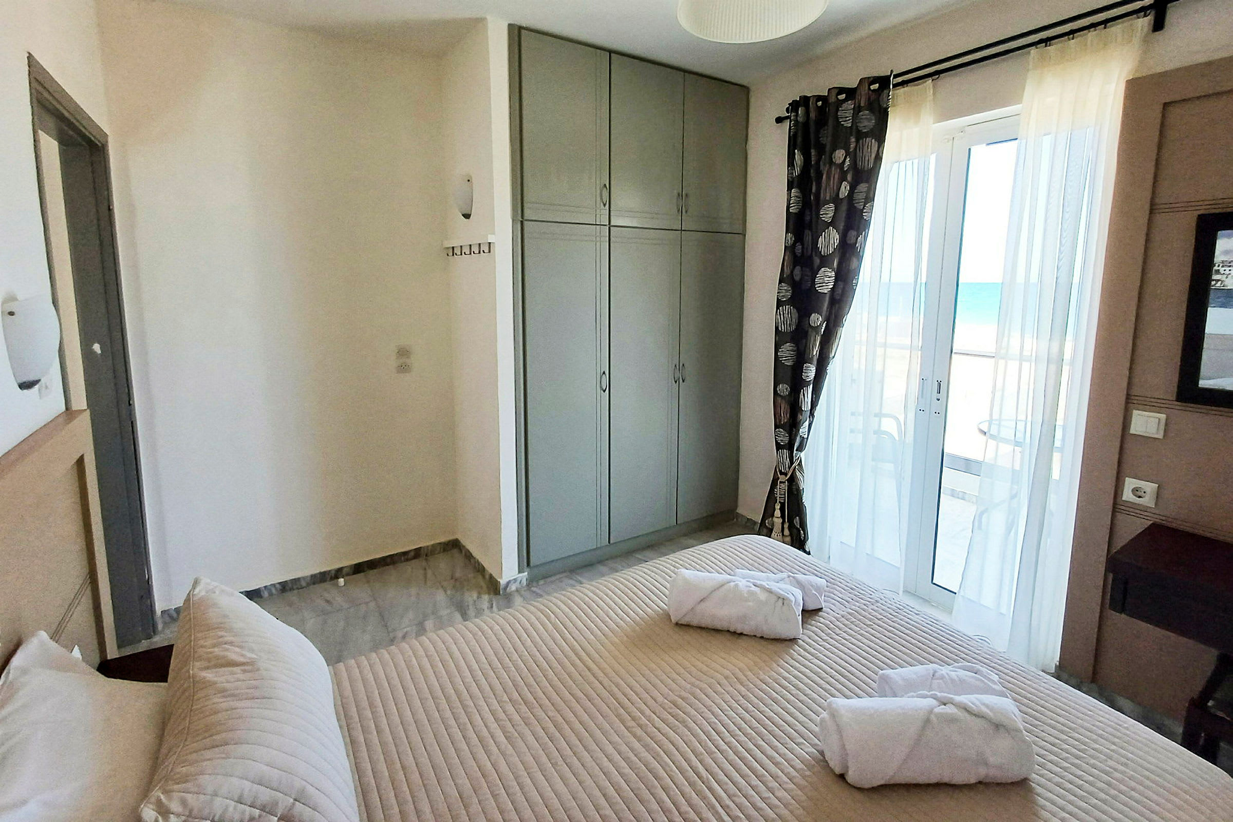 Ett hotellrum med en säng, grå garderob och en balkongdörr med havsutsikt. (AI-genererad text)