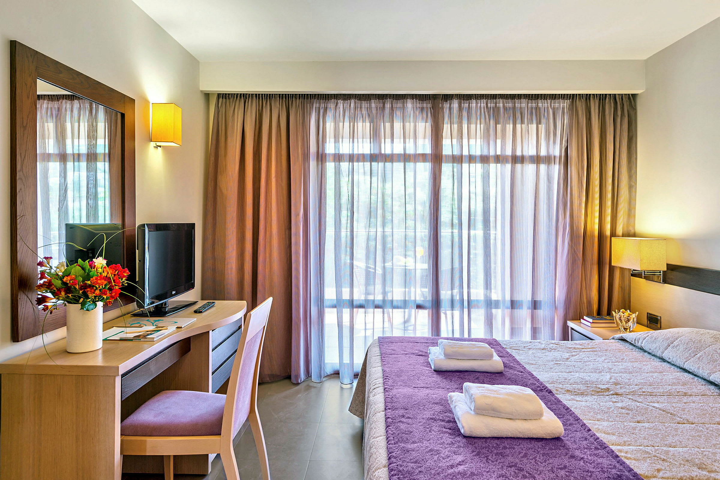 Ett hotellrum med en säng klädd i lila överkast, skrivbord med en TV och blombukett, samt ett stort fönster med genomskinliga gardiner. (AI-genererad text)