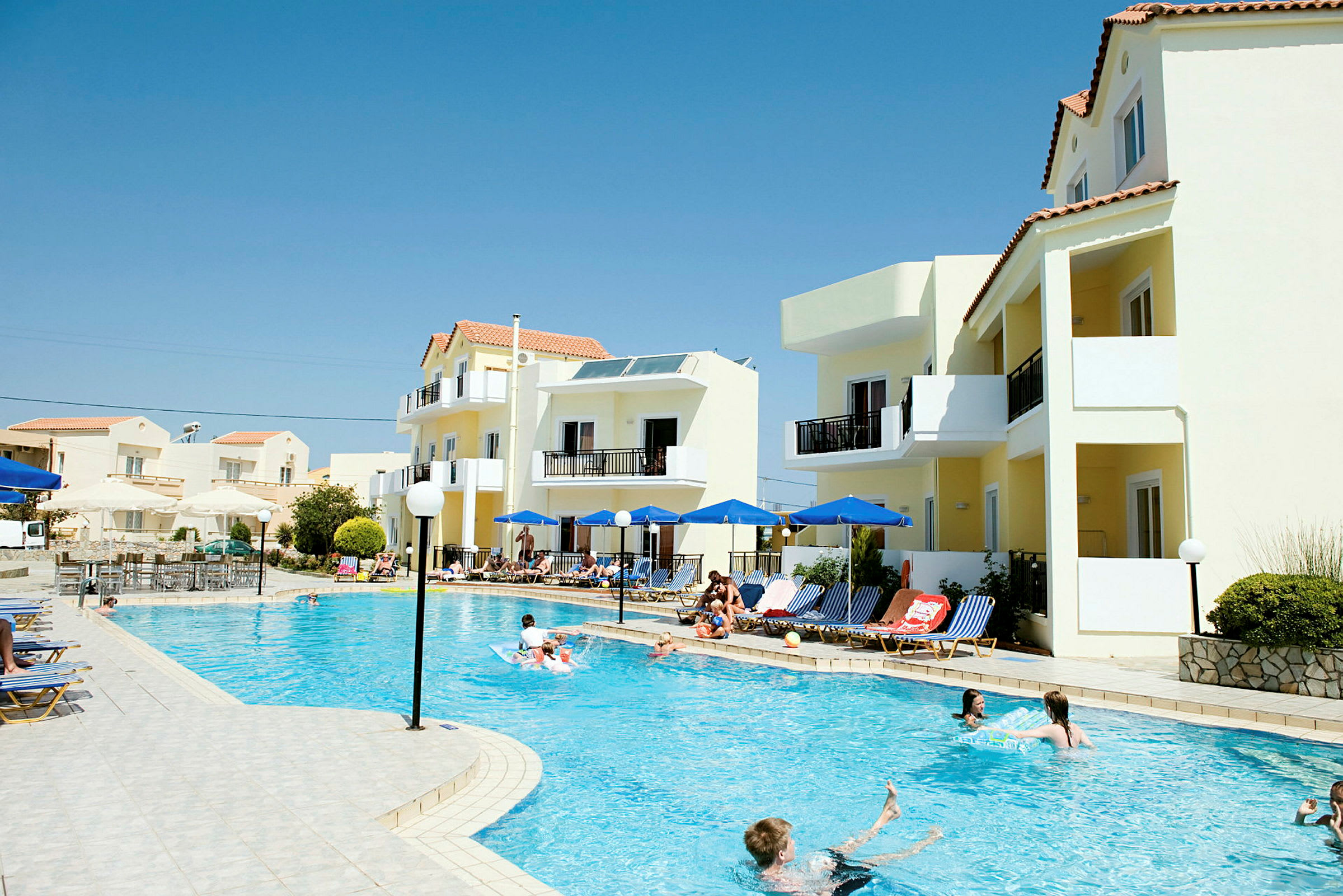 Utsikt över en pool med solstolar och parasoller vid sidan, omgiven av ljusa byggnader under en klarblå himmel. (AI-genererad text)