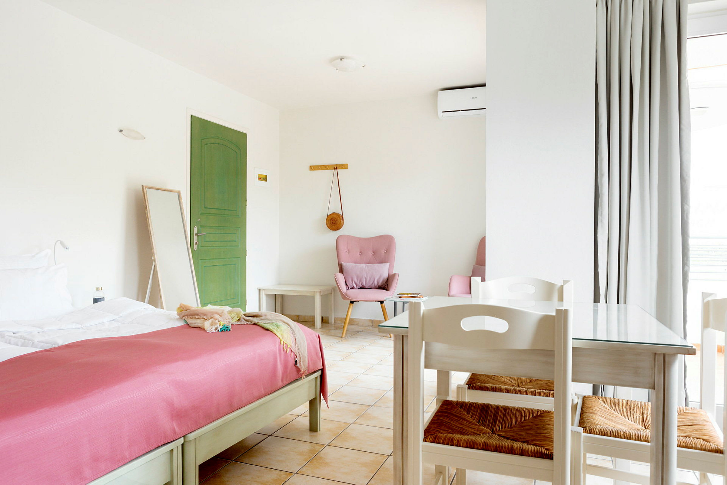 Ett ljus och rymligt hotellrum med en grön dörr, en säng med rosa överkast, och ett bord med stolar. (AI-genererad text)