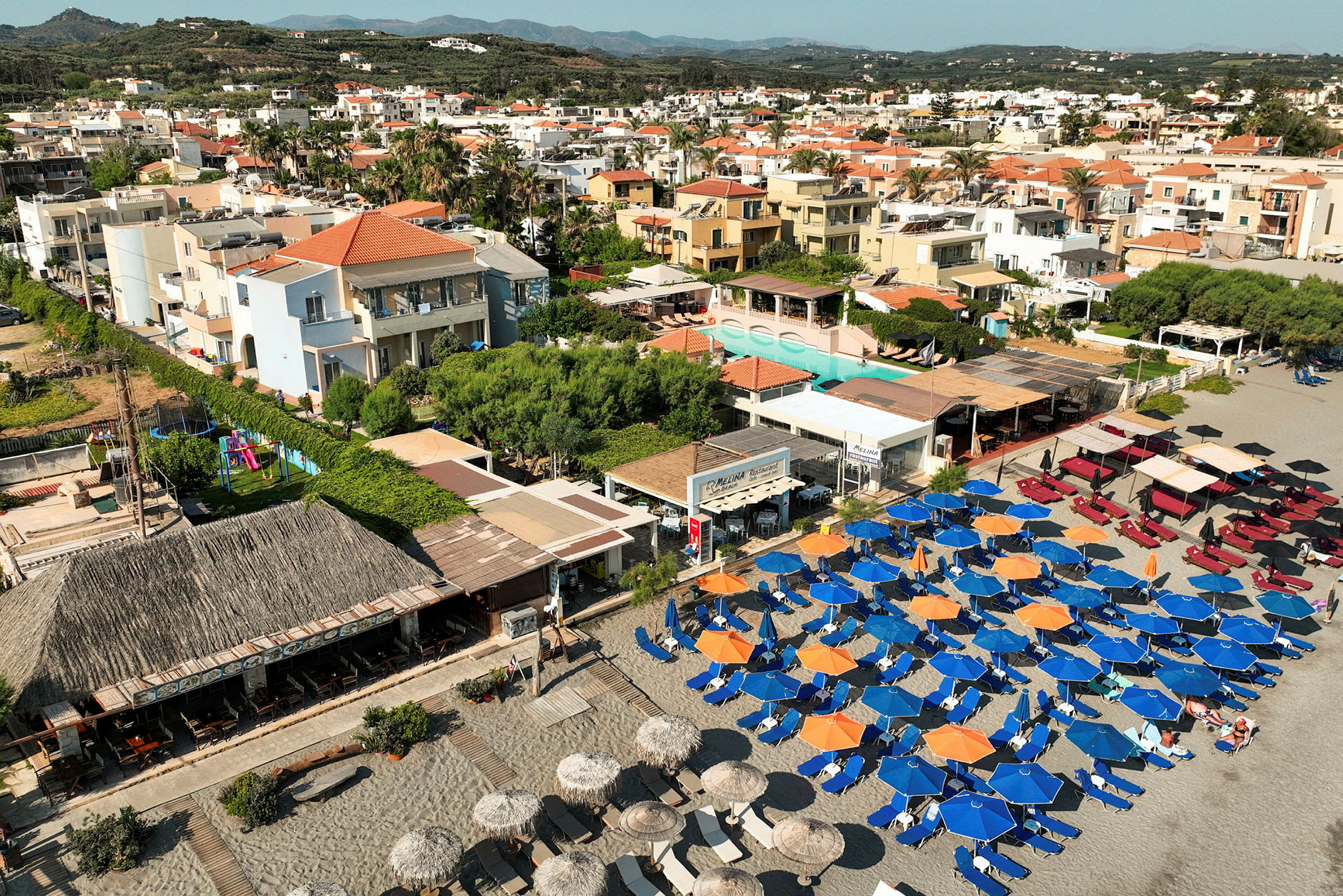 Flygfoto över stranden med solstolar och parasoller samt bostadsområden i bakgrunden. (AI-genererad text)