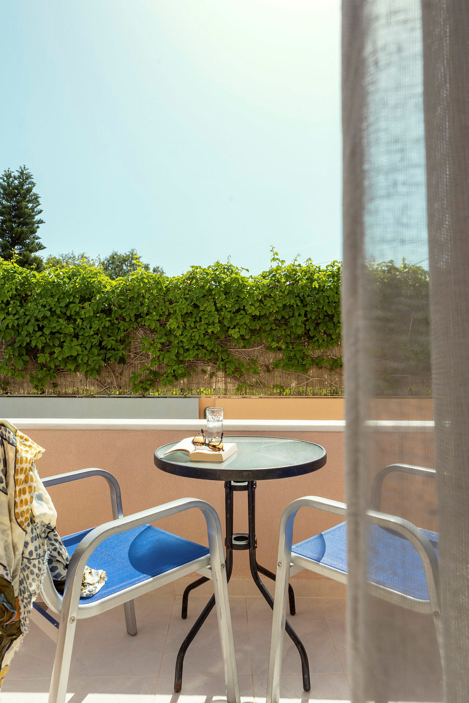 En balkong med två stolar och ett bord, där det finns ett glas, en bok och ett par solglasögon. Solen lyser och det är grönska i bakgrunden. (AI-genererad text)