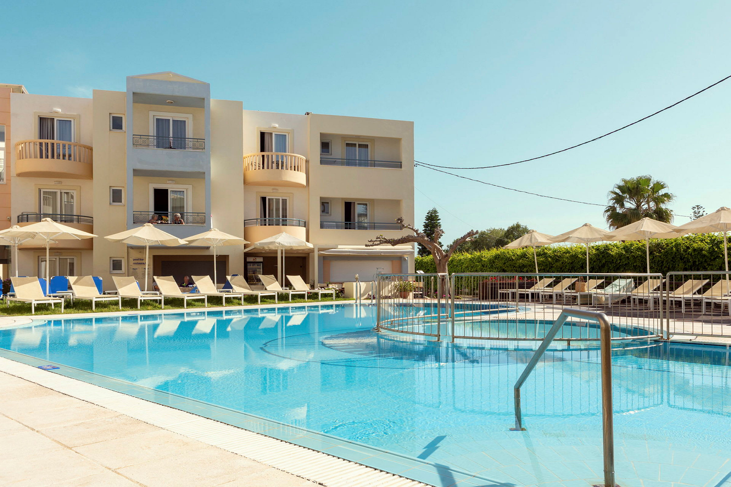 Bild av ett hotellområde med en pool, solstolar och parasoll. I bakgrunden syns en byggnad med balkonger. (AI-genererad text)
