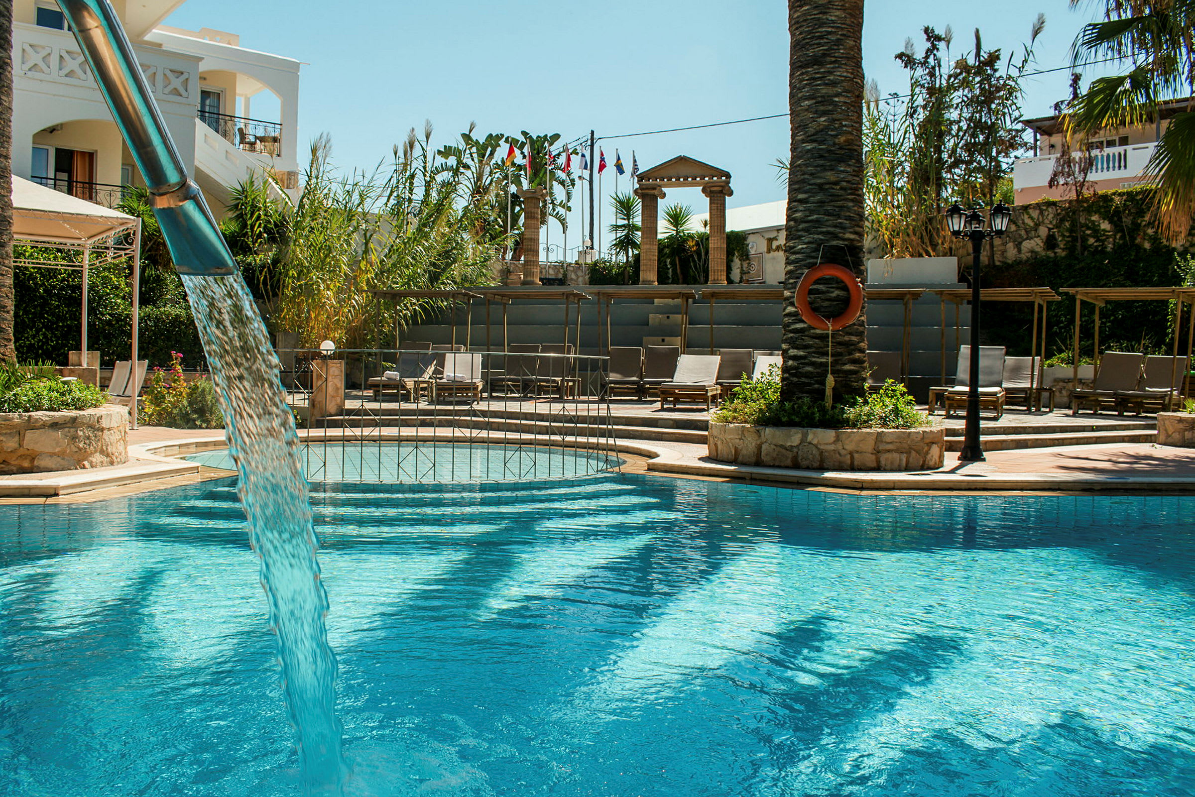En pool med klart vatten, omgiven av solstolar och grönska. Ett vattenfall rinner ner i poolen. (AI-genererad text)