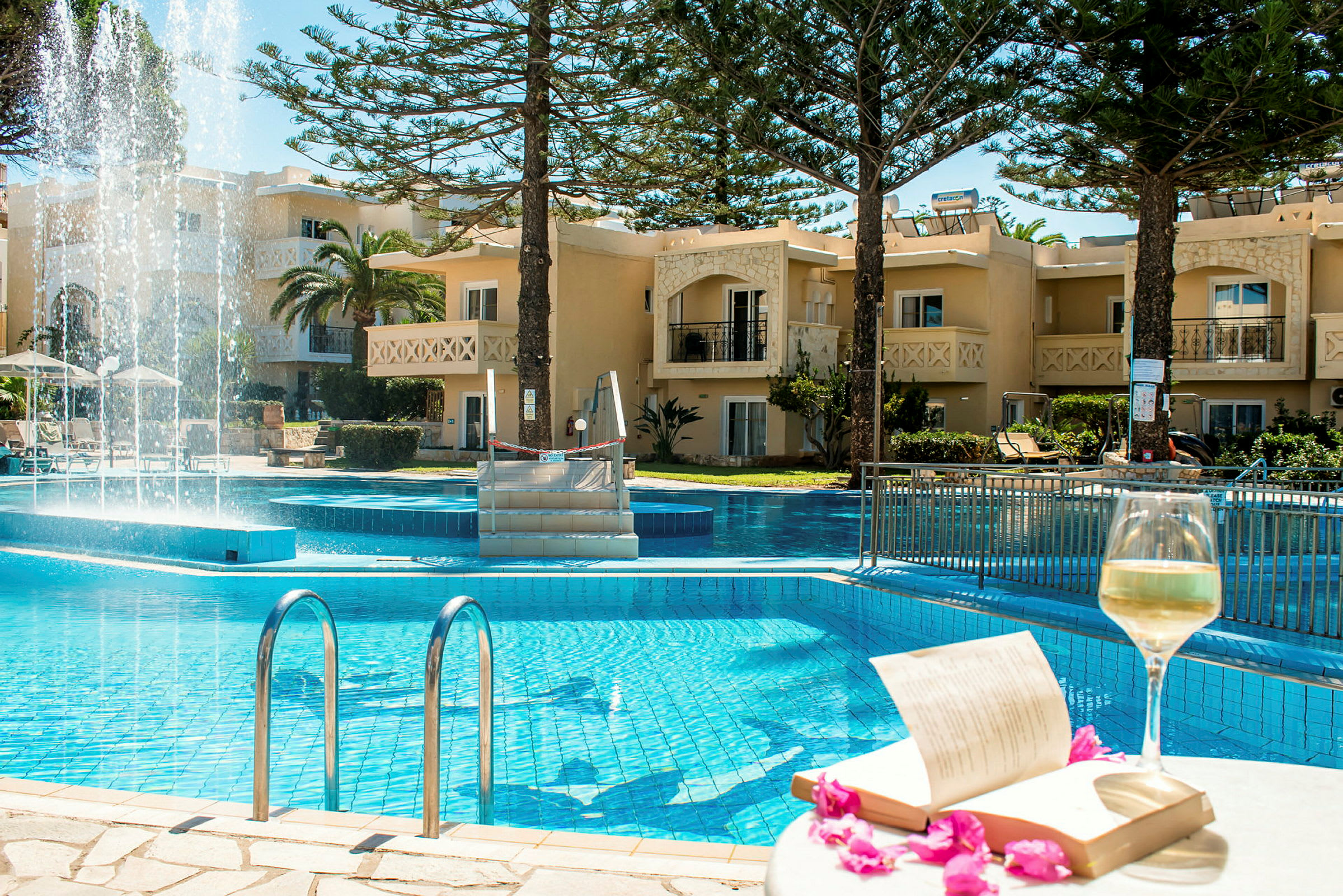 En pool med klart blått vatten framför en byggnad med balkonger och omgivande grönska. På en närliggande bordskant står ett glas med vitt vin bredvid en öppnad bok och några rosa blommor. (AI-genererad text)