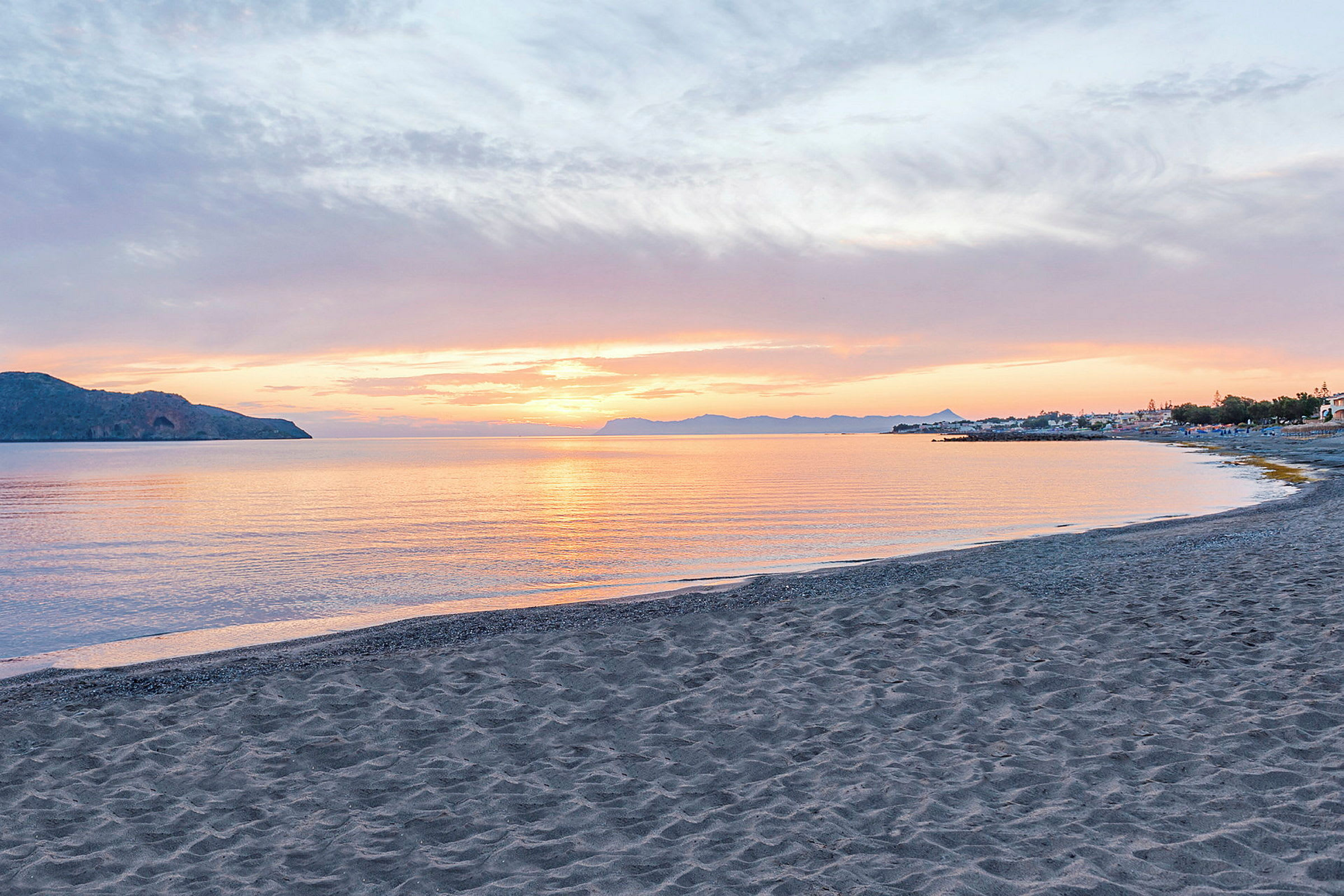 Solnedgång från en strand med havet i förgrunden och berg samt byggnader i bakgrunden. (AI-genererad text)