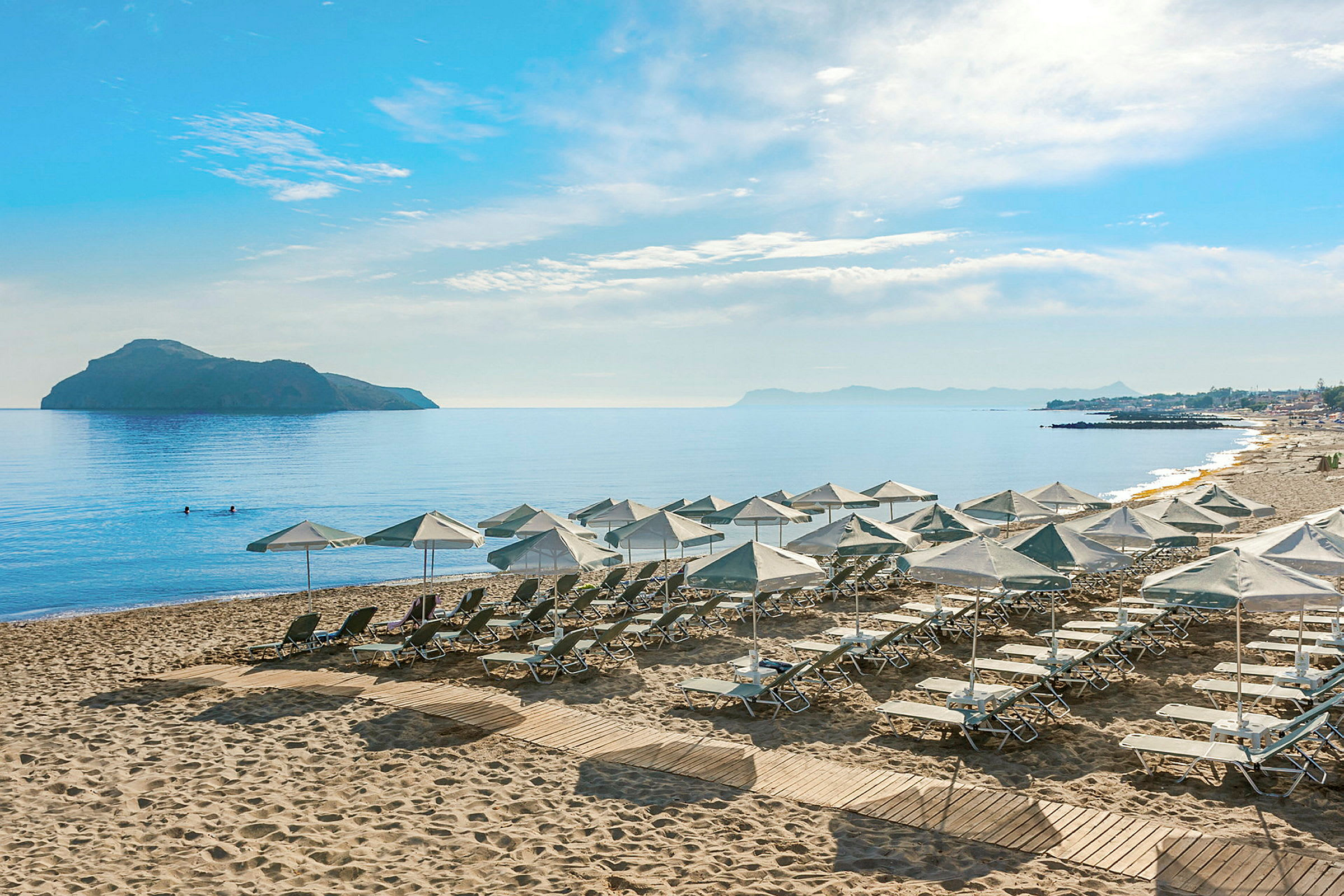 Utsikt över en strand med solstolar och parasoller, omgivande hav och en avlägsen ö. (AI-genererad text)