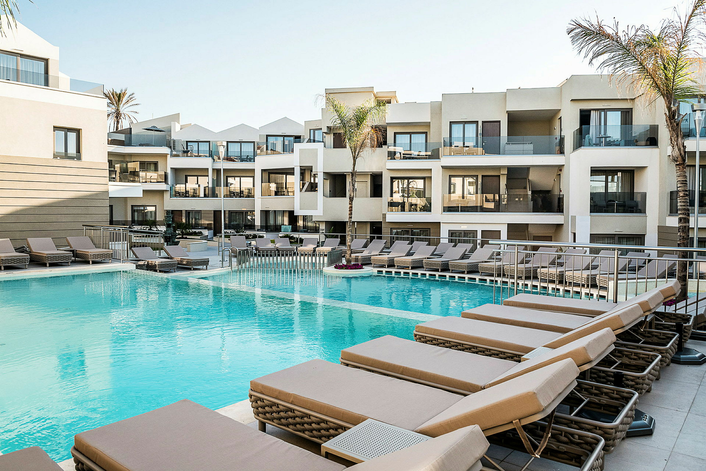 Hotellpool med solstolar och moderna byggnader i bakgrunden. (AI-genererad text)
