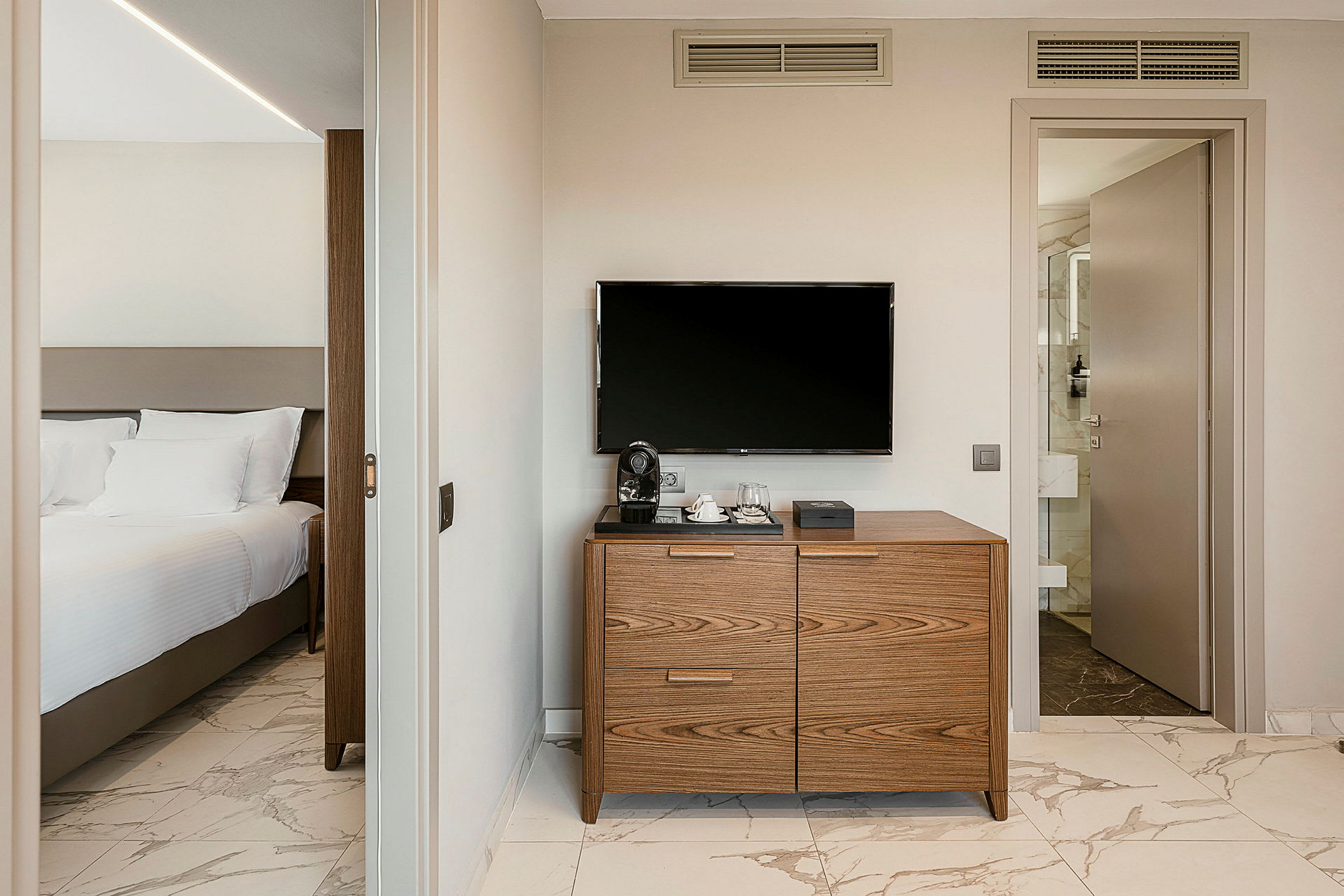 Ett hotellrum med en säng med vita sängkläder till vänster. En byrå med en kaffemaskin står under en TV på väggen till höger. (AI-genererad text)