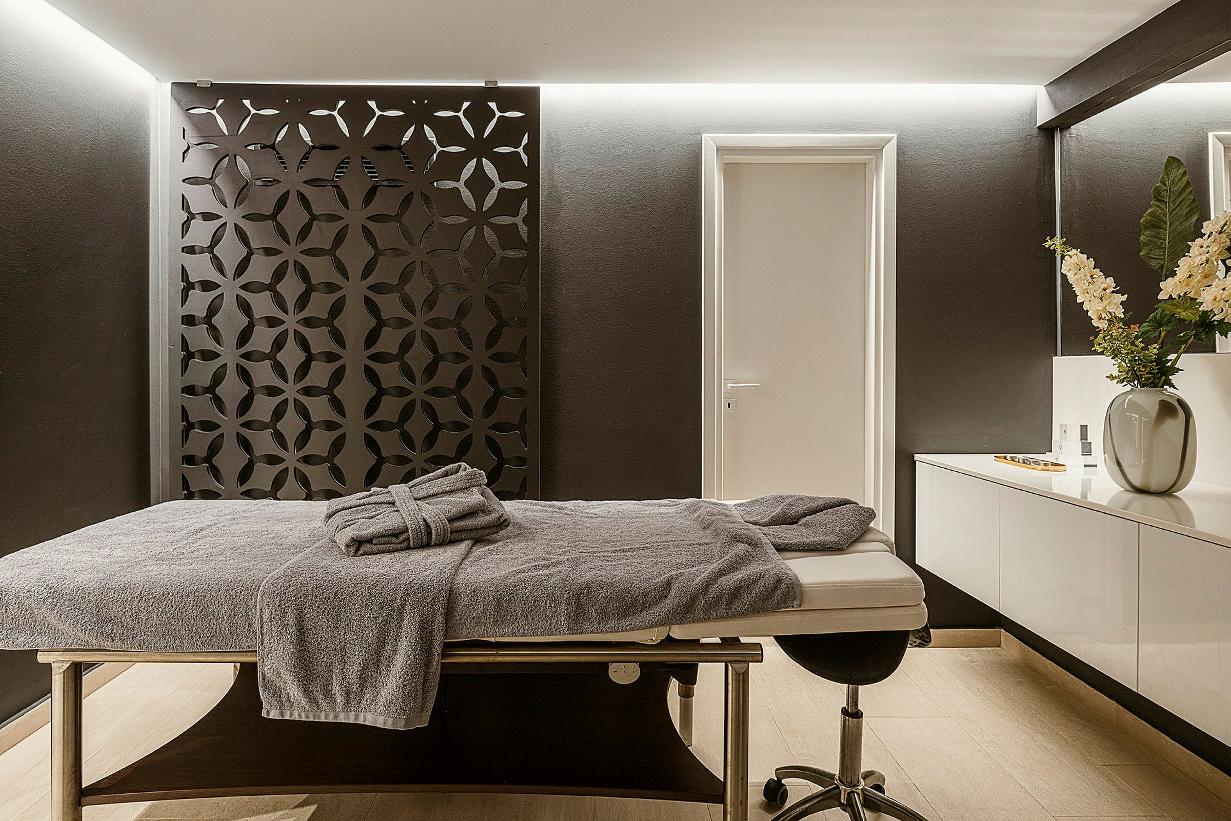 Ett rum med en massagebänk, handduk och spegel med blommor på en vit skänk. (AI-genererad text)