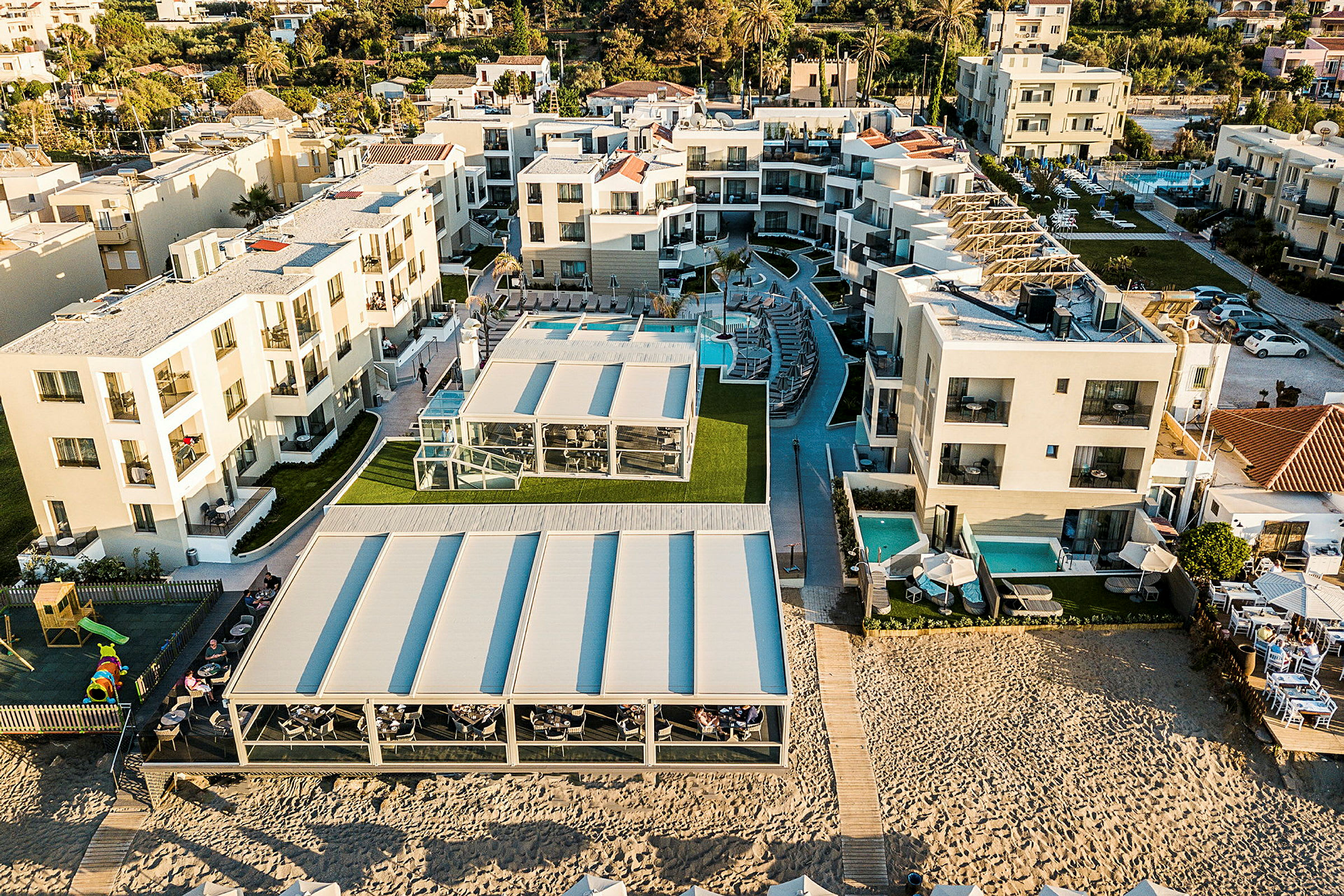 Flygbild över hotellområde med moderna byggnader, pooler och utomhusområden nära sandstrand. (AI-genererad text)