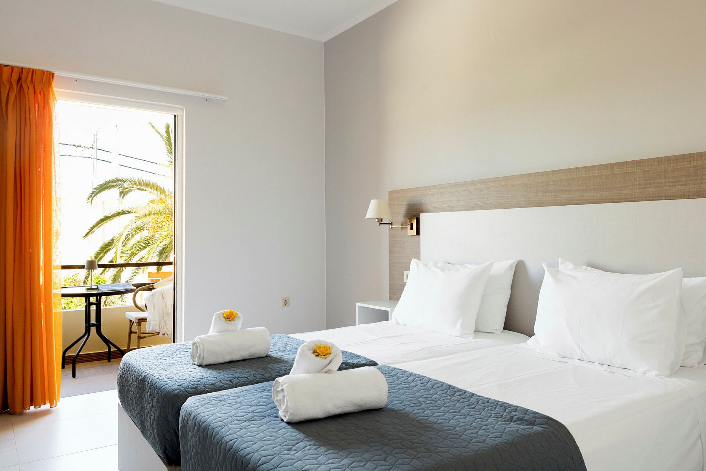 Ett hotellrum med två enkelsängar med grå överkast och vita kuddar samt handdukar dekorerade med gula blommor. Ett fönster med orange gardiner öppnar mot en balkong med utsikt över palmer. (AI-genererad text)