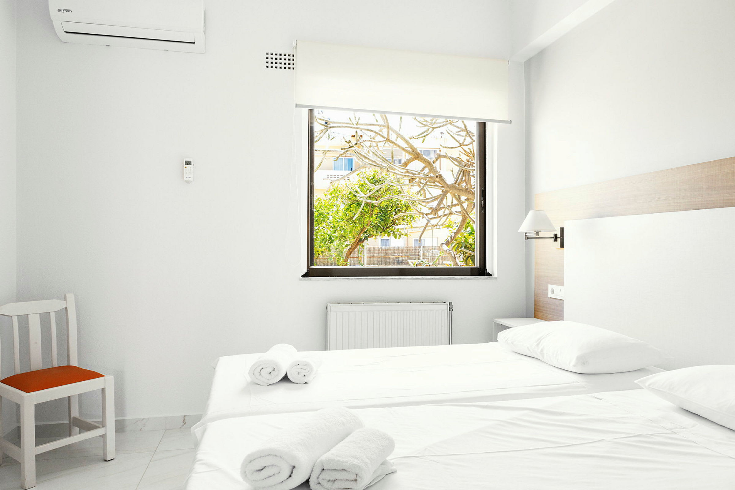 Ett ljust hotellrum med en bäddad säng, vita väggar och en stol med orange sits. Ett fönster med utsikt över träd och en byggnad utanför. (AI-genererad text)