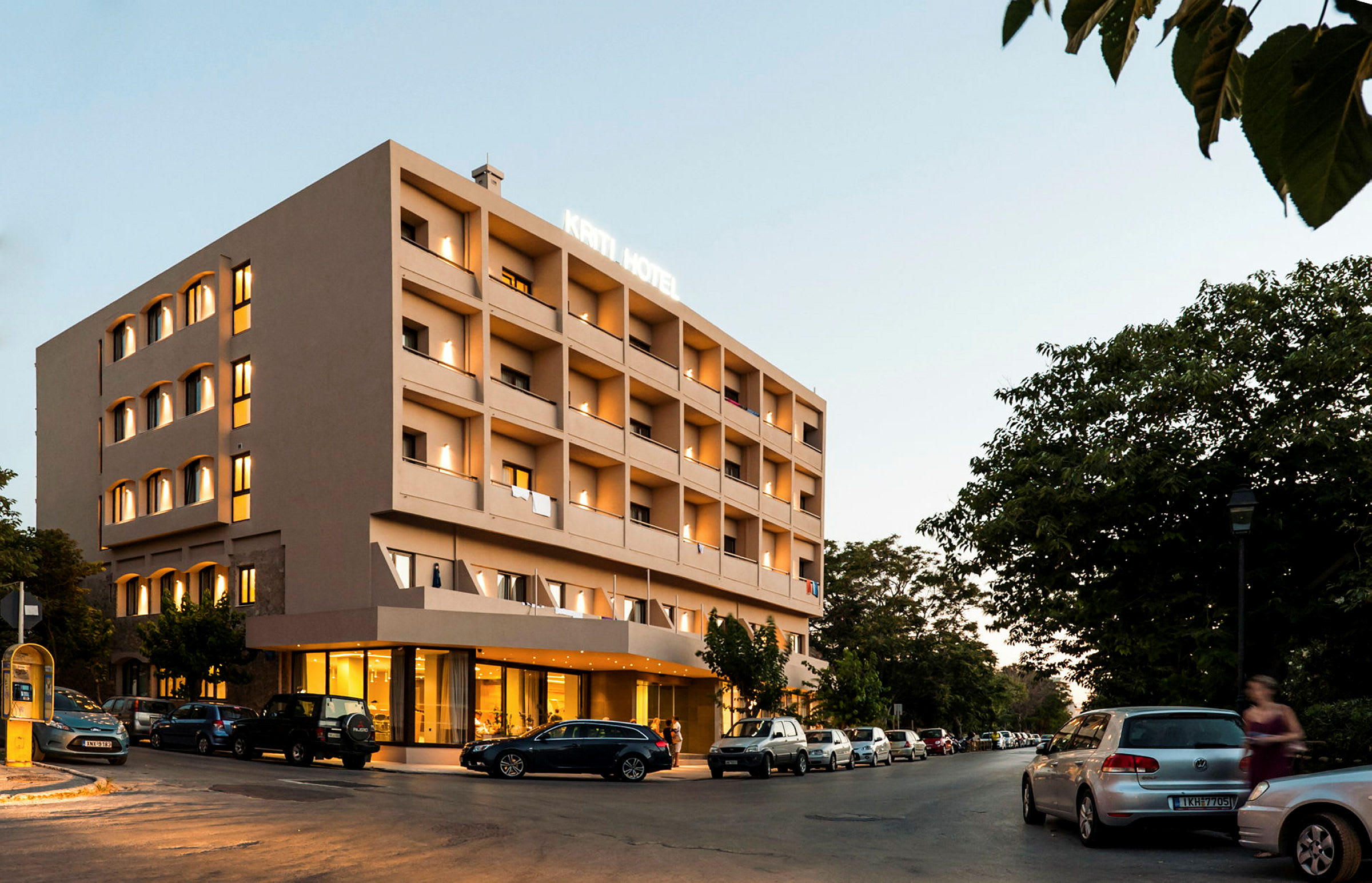 Fyravånings hotellbyggnad med belyst fasad, parkerade bilar runtom och gröna träd i bakgrunden. (AI-genererad text)