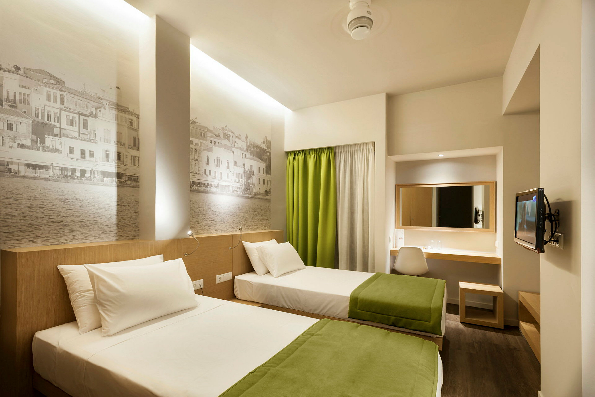 Två enkelsängar med gröna filtar i ett modernt hotellrum med väggmålningar och skrivbord. (AI-genererad text)
