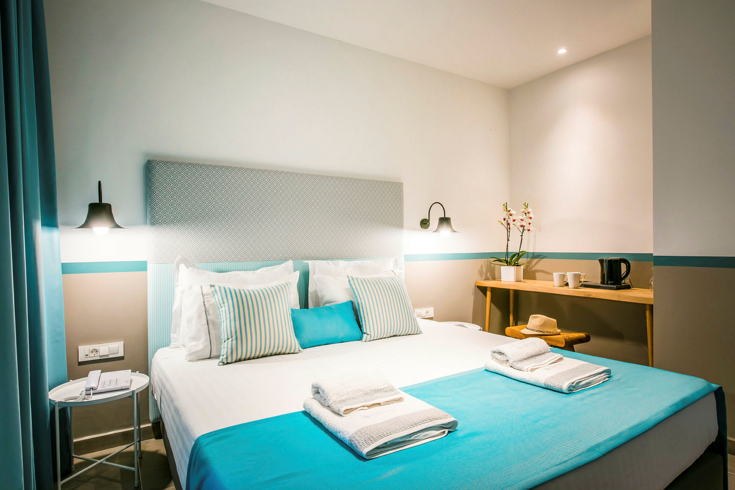 Ljust och modernt hotellrum med en dubbelsäng bäddad med blå och vita detaljer. På sängen finns handdukar, och ett sidobord med lampa samt ett skrivbord med dekorativ blomma och vattenkokare visas. (AI-genererad text)