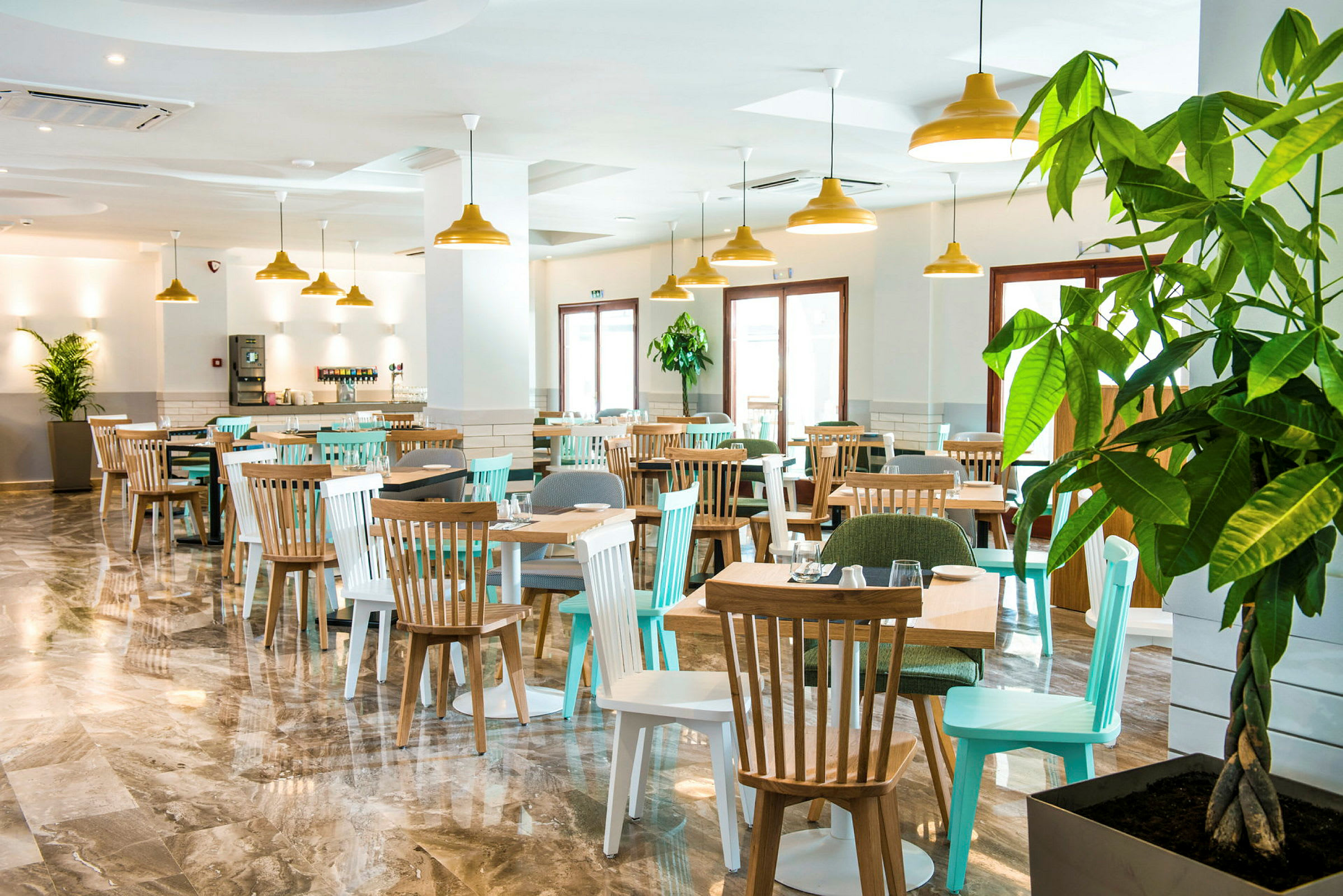 En restaurang med bord och stolar samt stora fönster och gröna växter. (AI-genererad text)