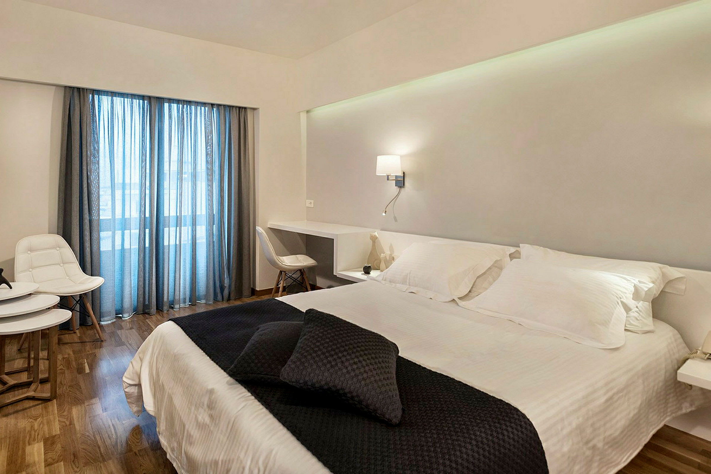 Ett hotellrum med en dubbelsäng klädd i vita lakan och mörka prydnadskuddar. Rummet har ett skrivbord, vita möbler och glasdörrar med blåa gardiner. (AI-genererad text)