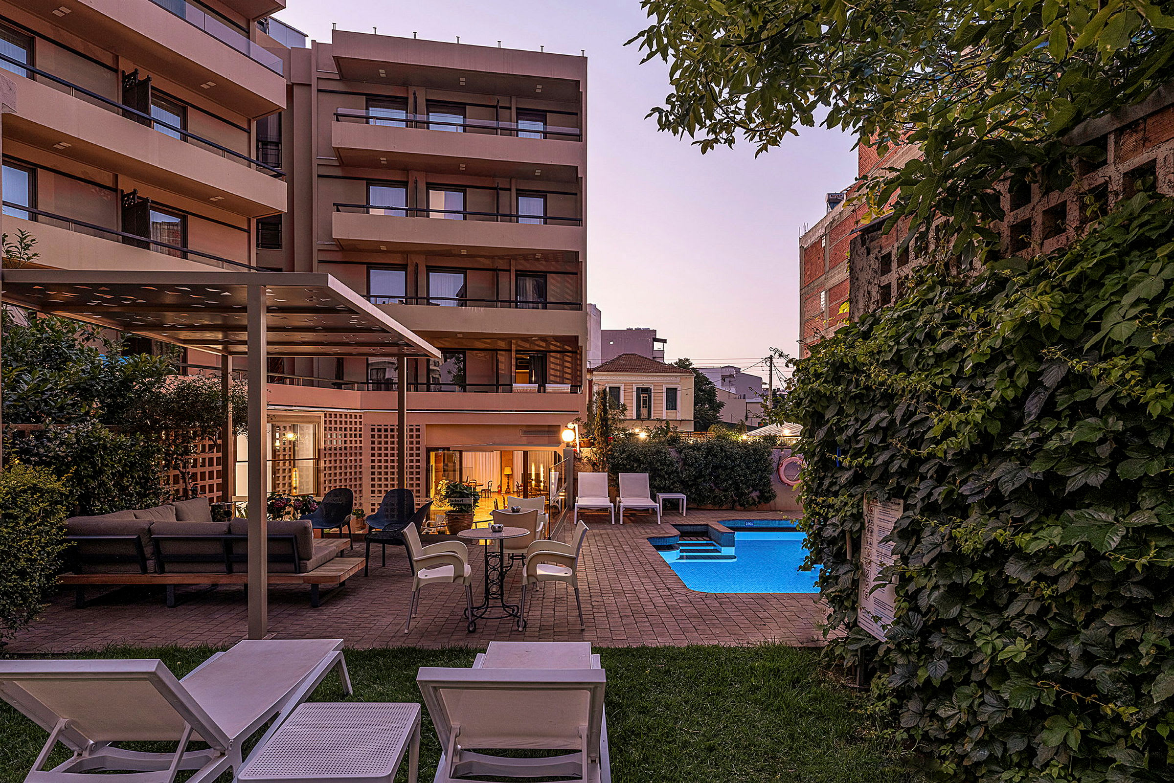 Bild av en utomhusmiljö med solstolar, sittmöbler, en swimmingpool och grönska framför en byggnad i kvällsljus. (AI-genererad text)