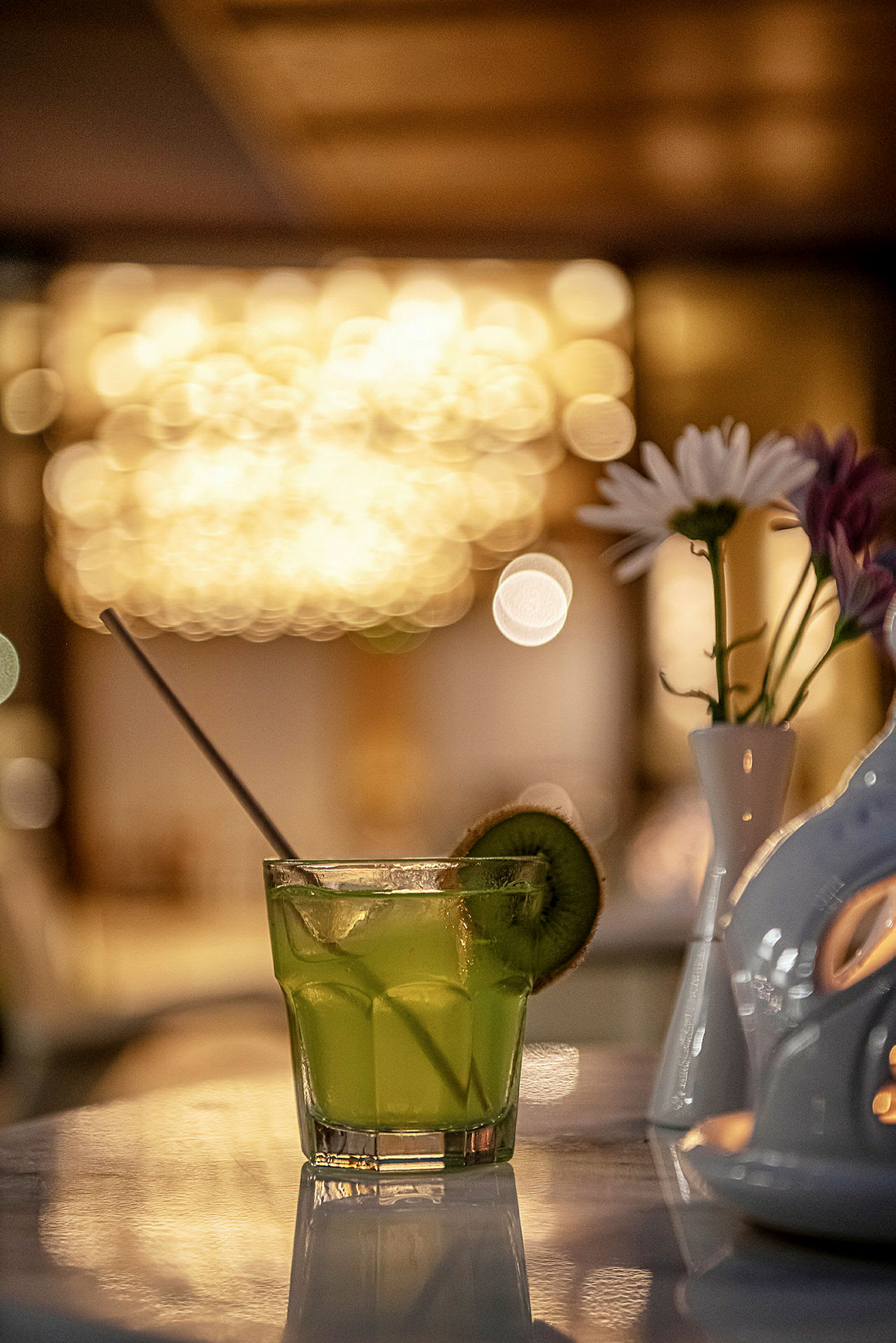 Ett glas med en grön drink dekorerad med en kiwiskiva på en marmorbord med en vas som innehåller blommor i bakgrunden. (AI-genererad text)