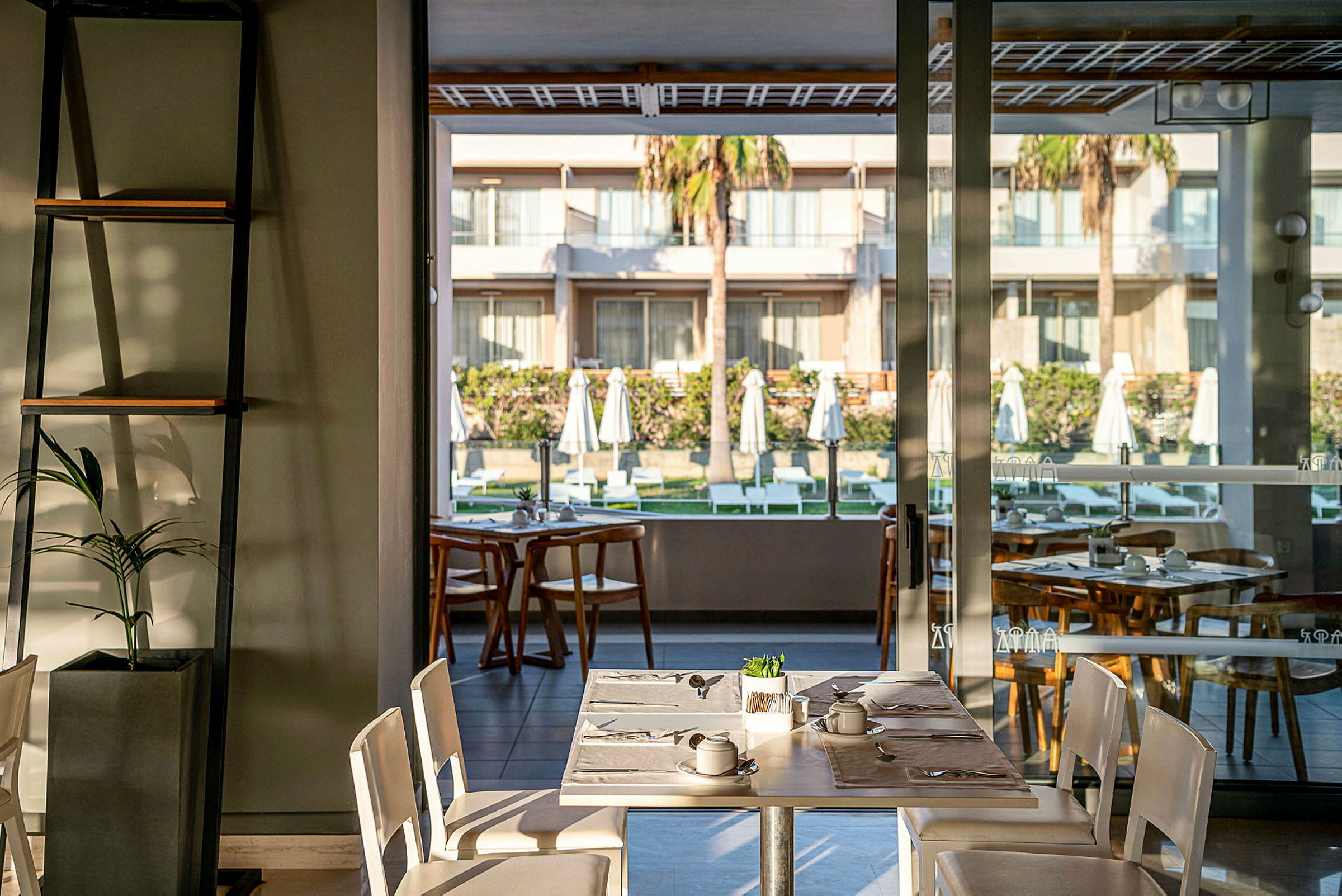 En restaurang med bord i förgrunden och utsikt över en innergård med solstolar och parasoller. (AI-genererad text)