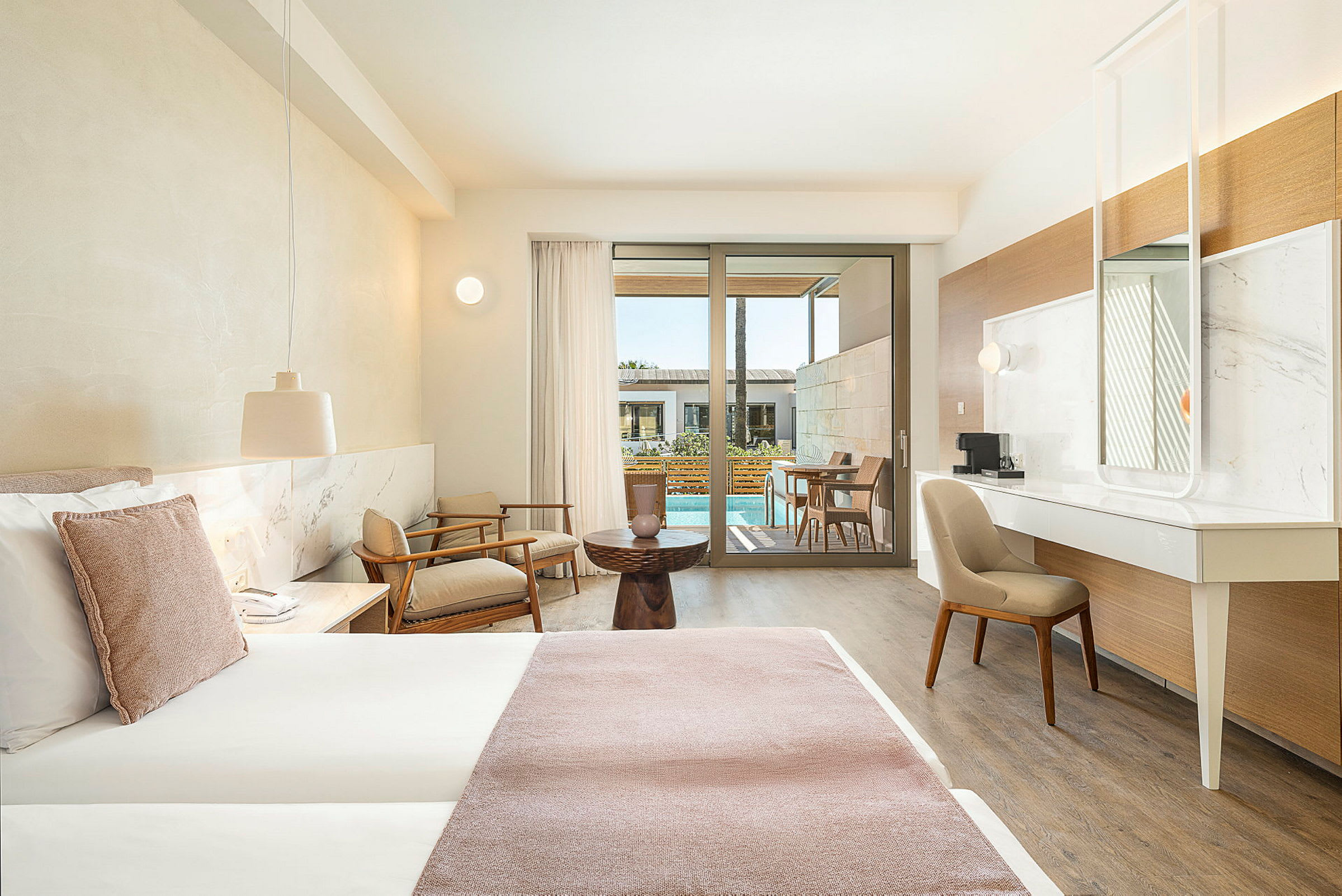 Ett hotellrum med modern inredning, en stor säng, sittområde och utsikt mot en terrass med pool. (AI-genererad text)