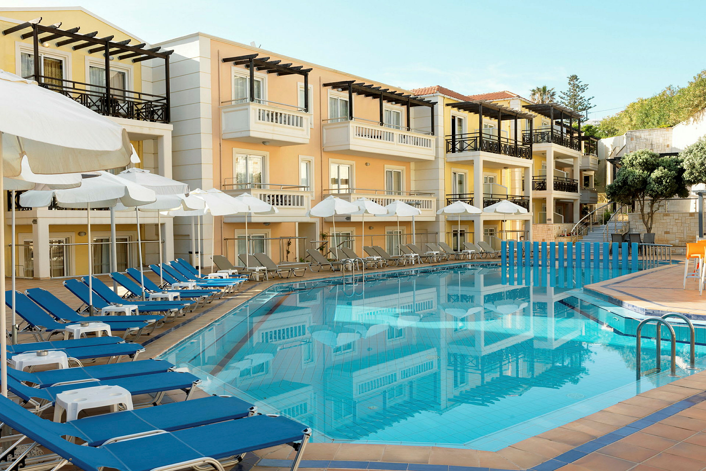 Hotellfasad i pastellfärger med balkonger, framför en klarblå swimmingpool omgiven av solstolar och parasoller. (AI-genererad text)