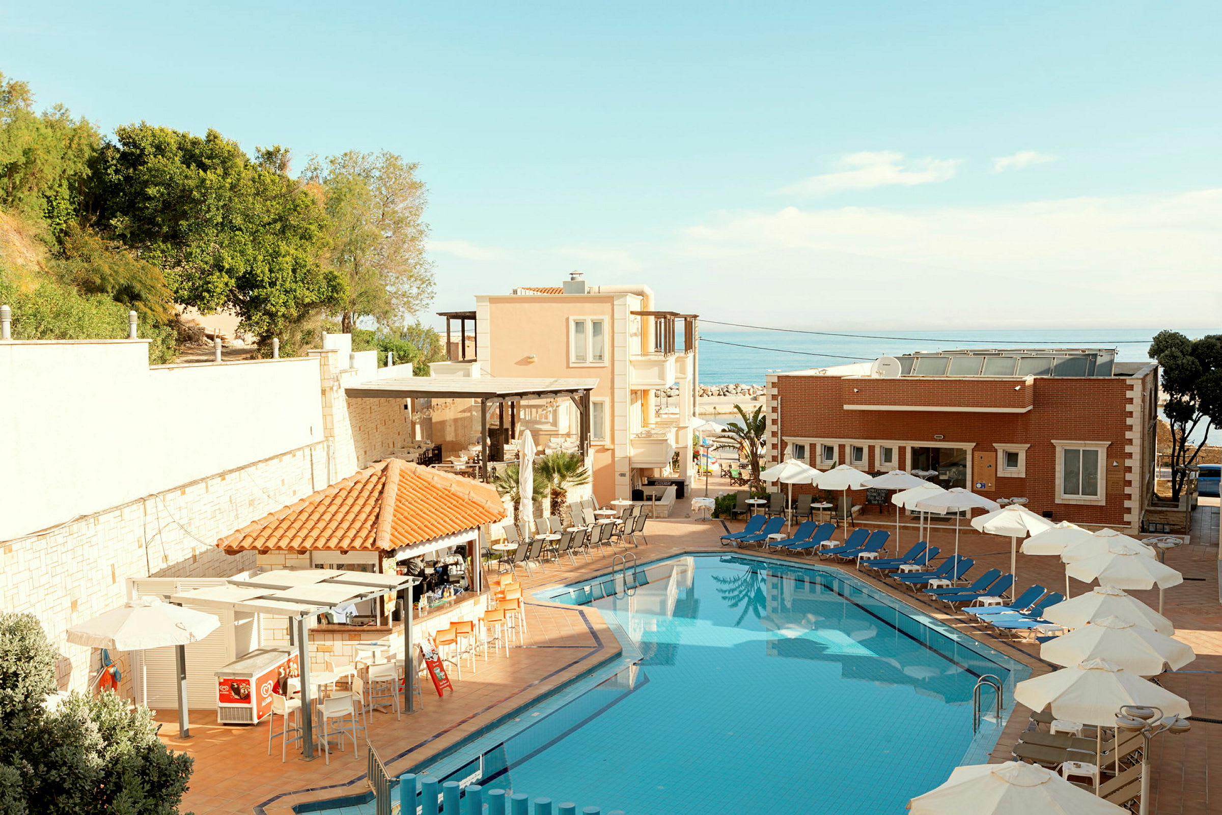 En hotellpool med solstolar och parasoller vid sidan om samt byggnader och grönska i bakgrunden. (AI-genererad text)